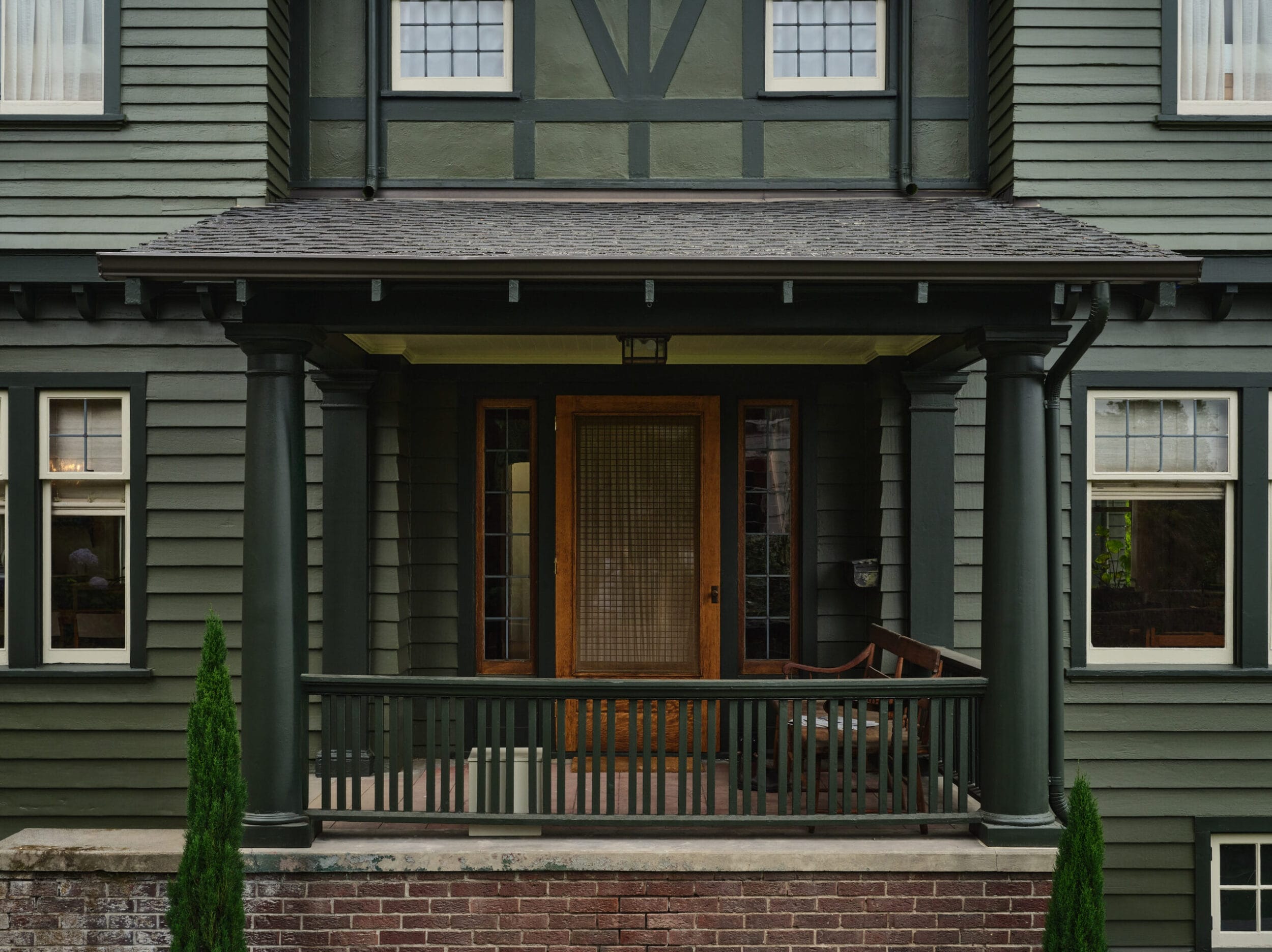 Two-story green house with a covered front porch, wooden door, and windows. Porch has two columns and a bench. Brick base and small plants in front.