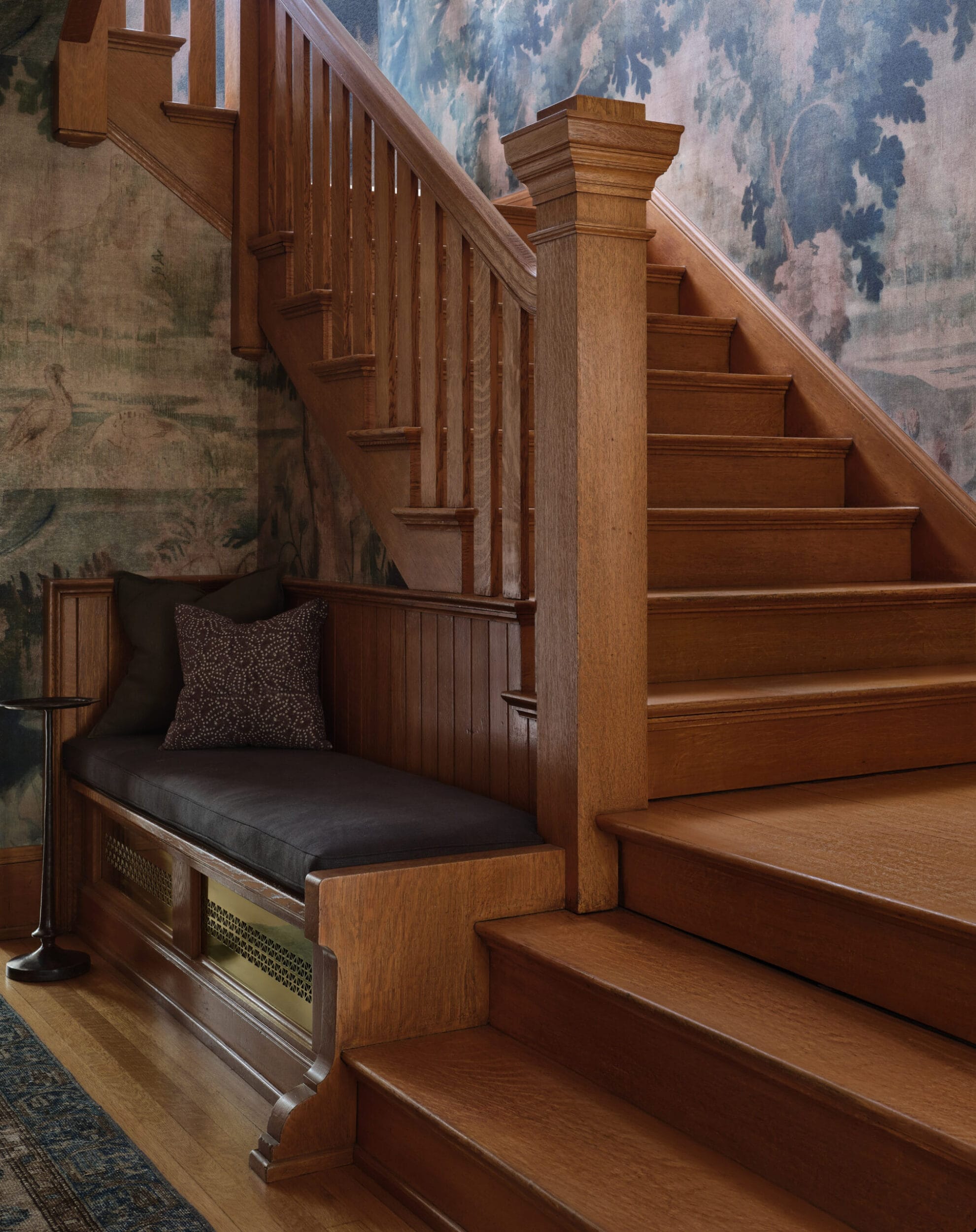 Wooden staircase with a built-in bench featuring dark cushions and a decorative pillow, set against a wall with a scenic mural.