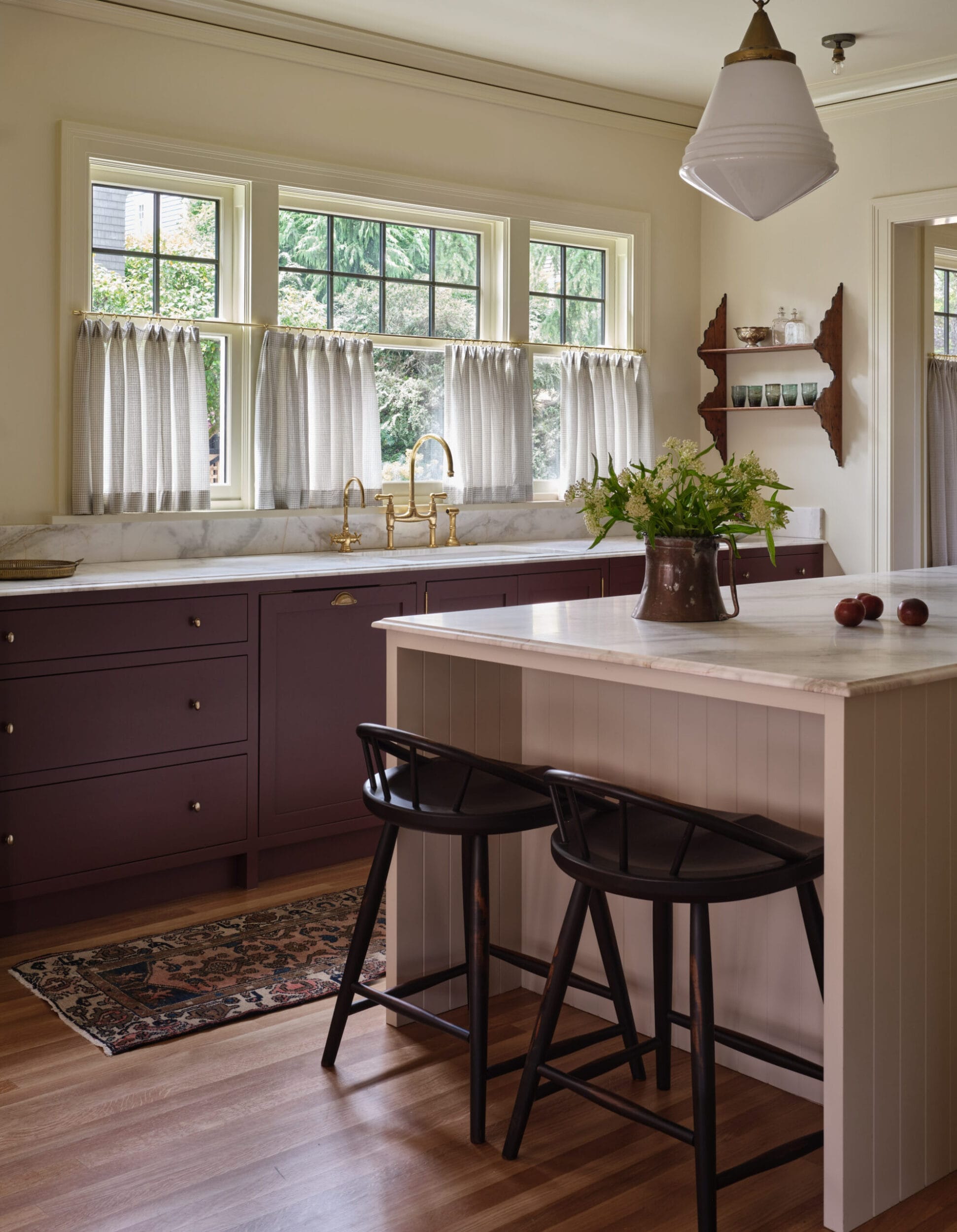 A cozy kitchen with a marble-topped island, dual black stools, and maroon cabinets. Curtains cover the sunny windows, and a vase with flowers sits on the island alongside a few apples.