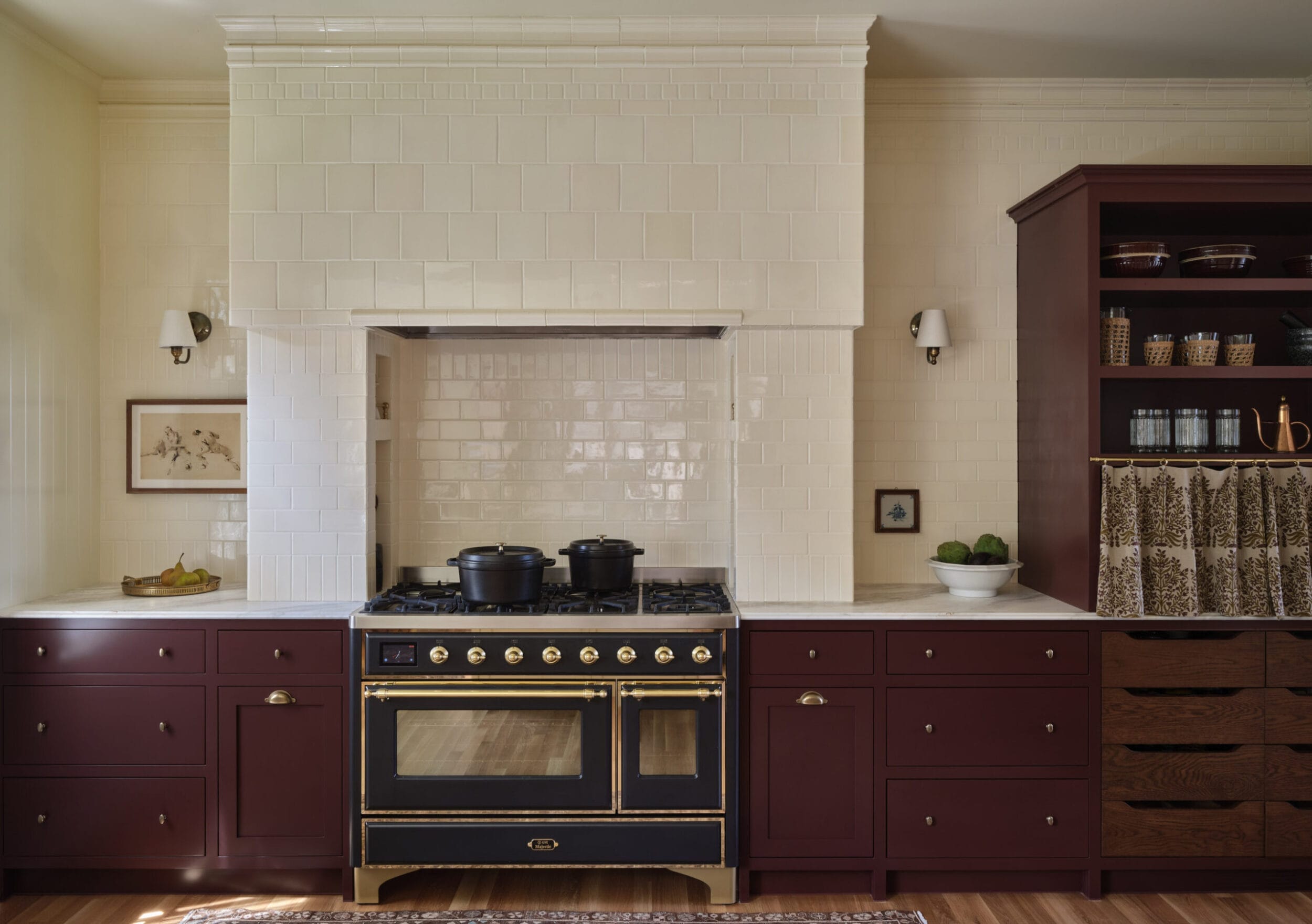 A kitchen features a vintage-style black and gold stove with two pots. It is flanked by maroon cabinets and white tiles. A bowl of green apples and a framed picture are on the counter.