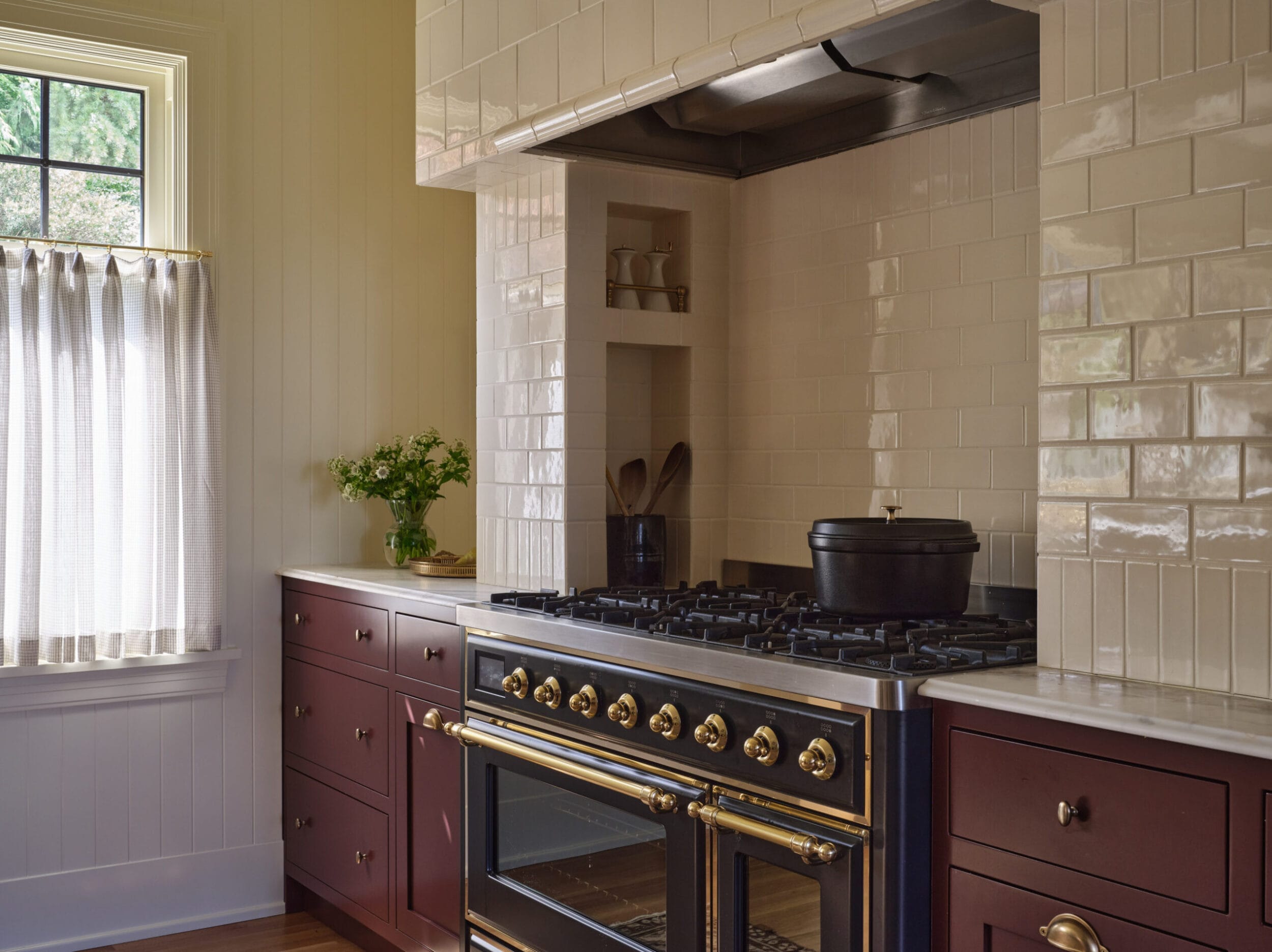 A kitchen with a maroon stove and oven, beige tiled backsplash, wooden cabinets, a window with curtains, and a pot on the stove.