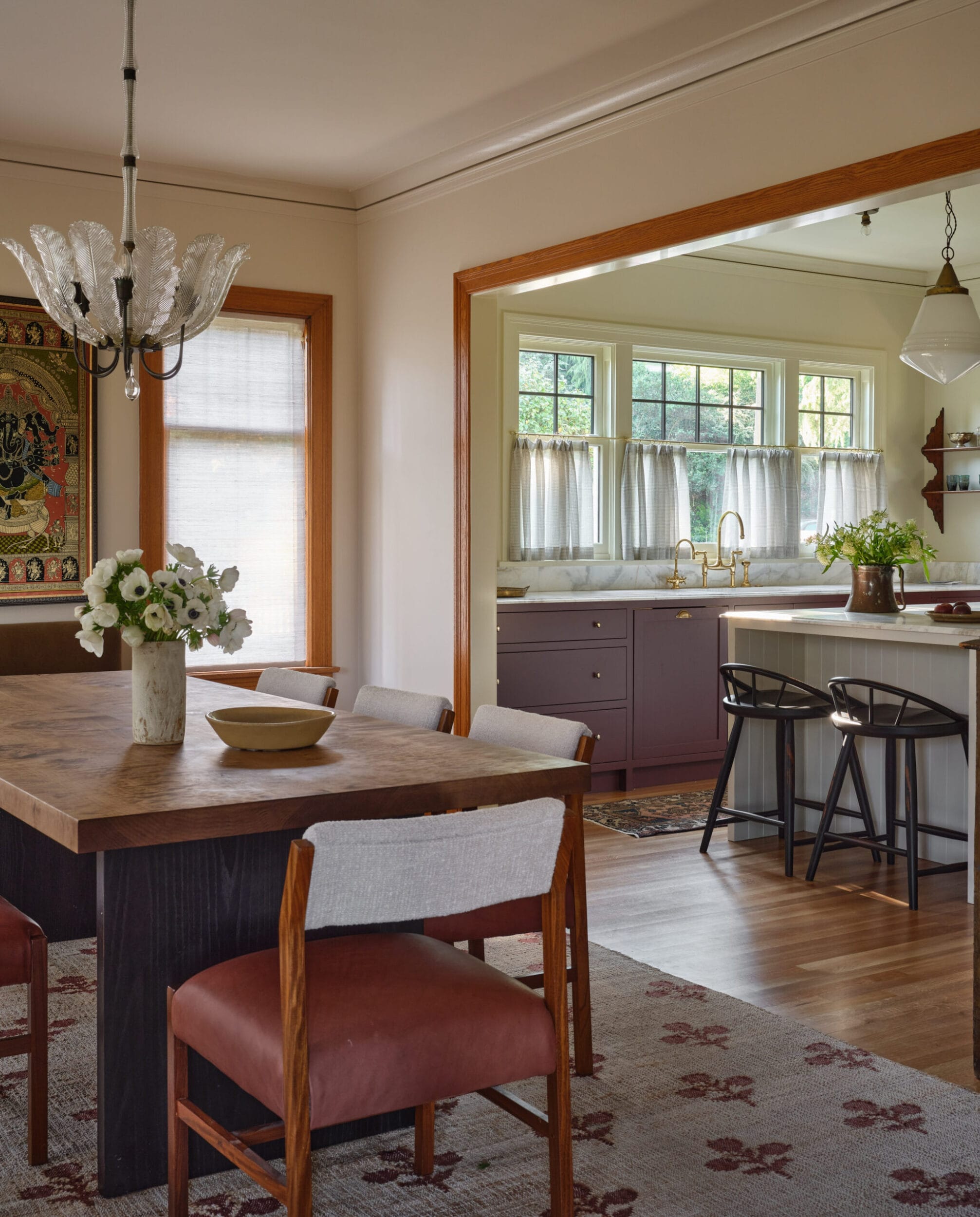 Dining room with a wooden table, cushioned chairs, and a floral centerpiece. Adjacent kitchen features purple cabinets, a white island with stools, and window curtains. Chandelier above table.