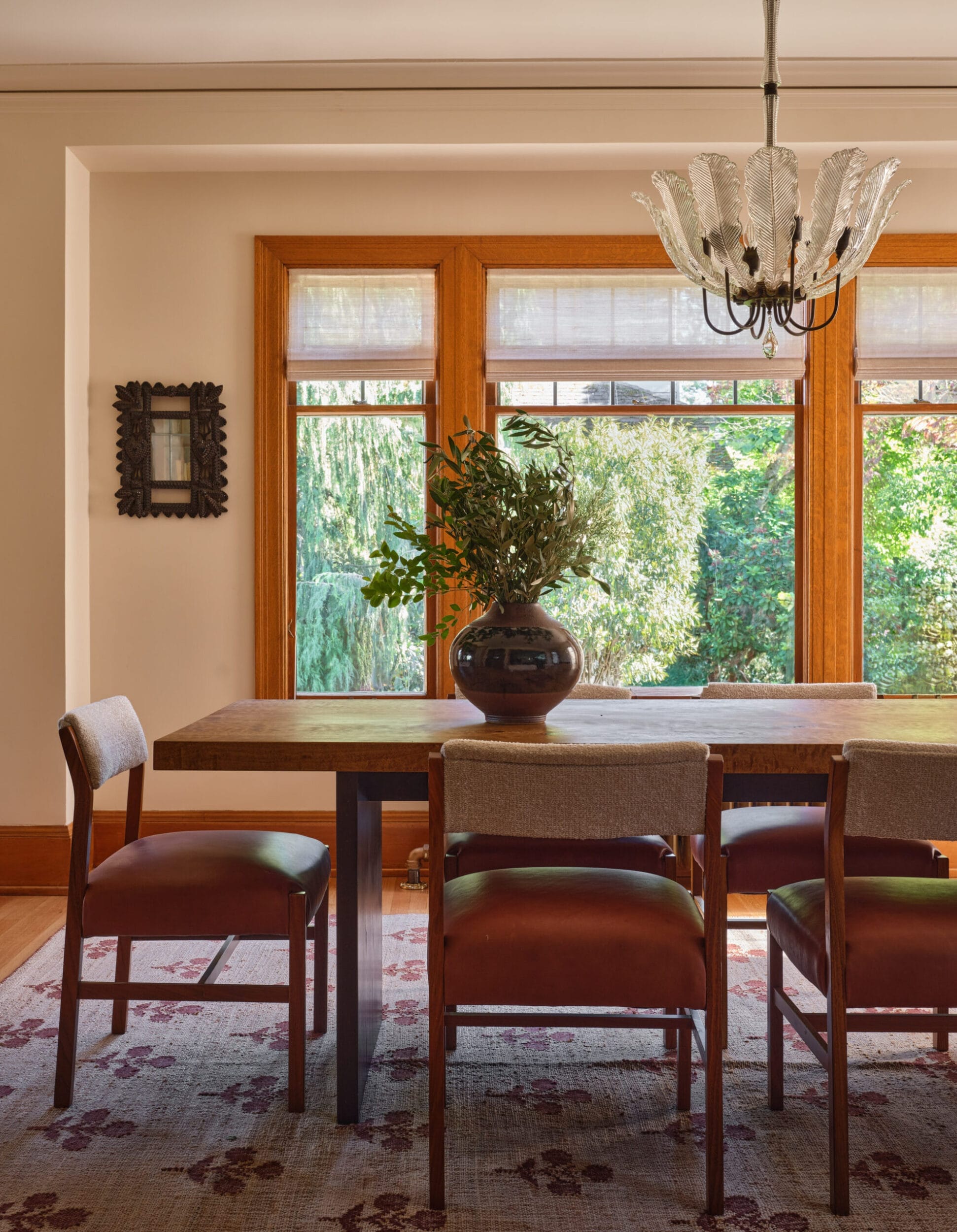 A dining room with a wooden table, four chairs, a vase with greenery, a decorative rug, and a chandelier. Large windows provide a view of greenery outside.