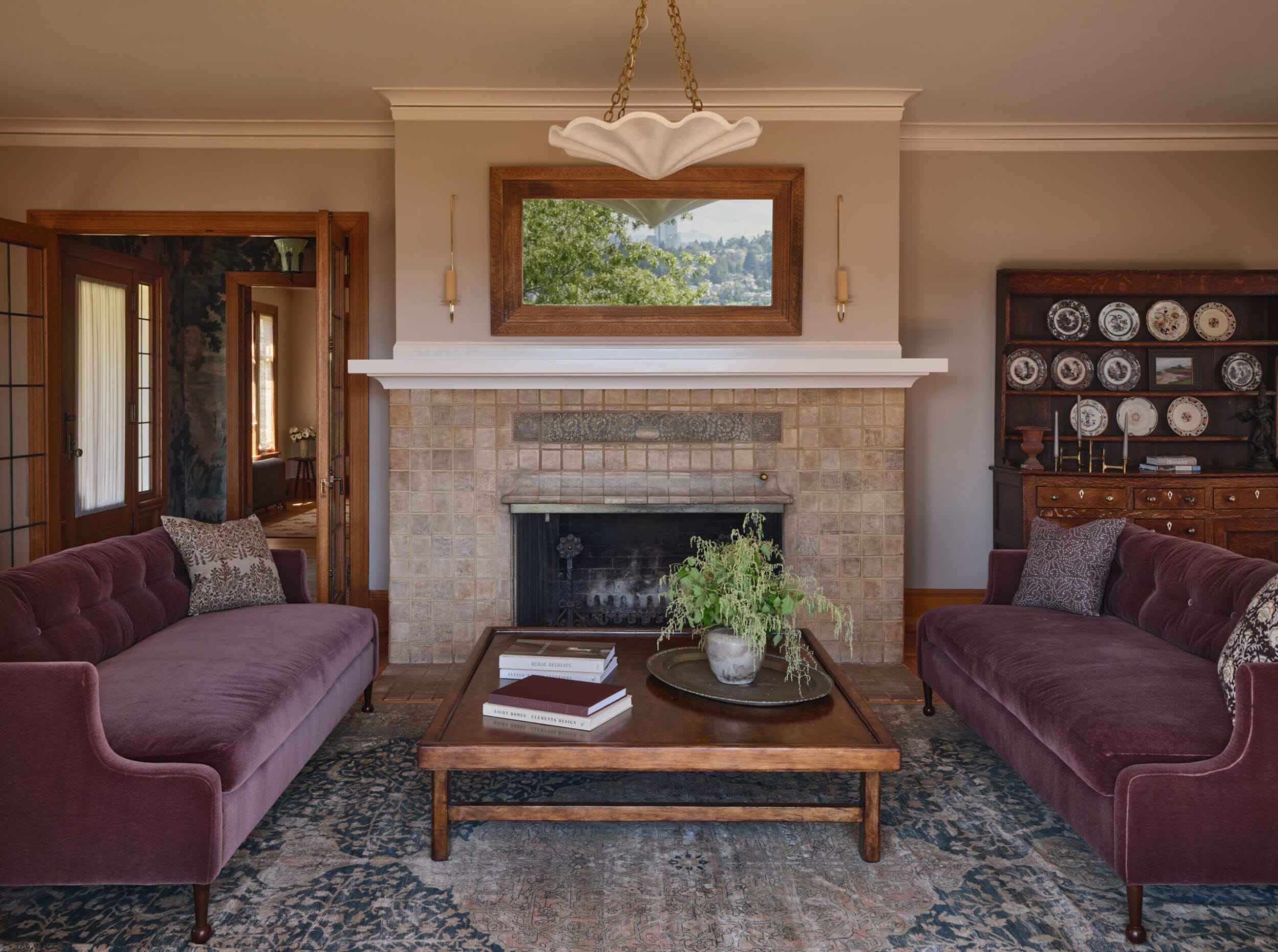 A living room with two velvet sofas, a coffee table, fireplace, and a wall mirror. A china cabinet and decorative plates are in the background.