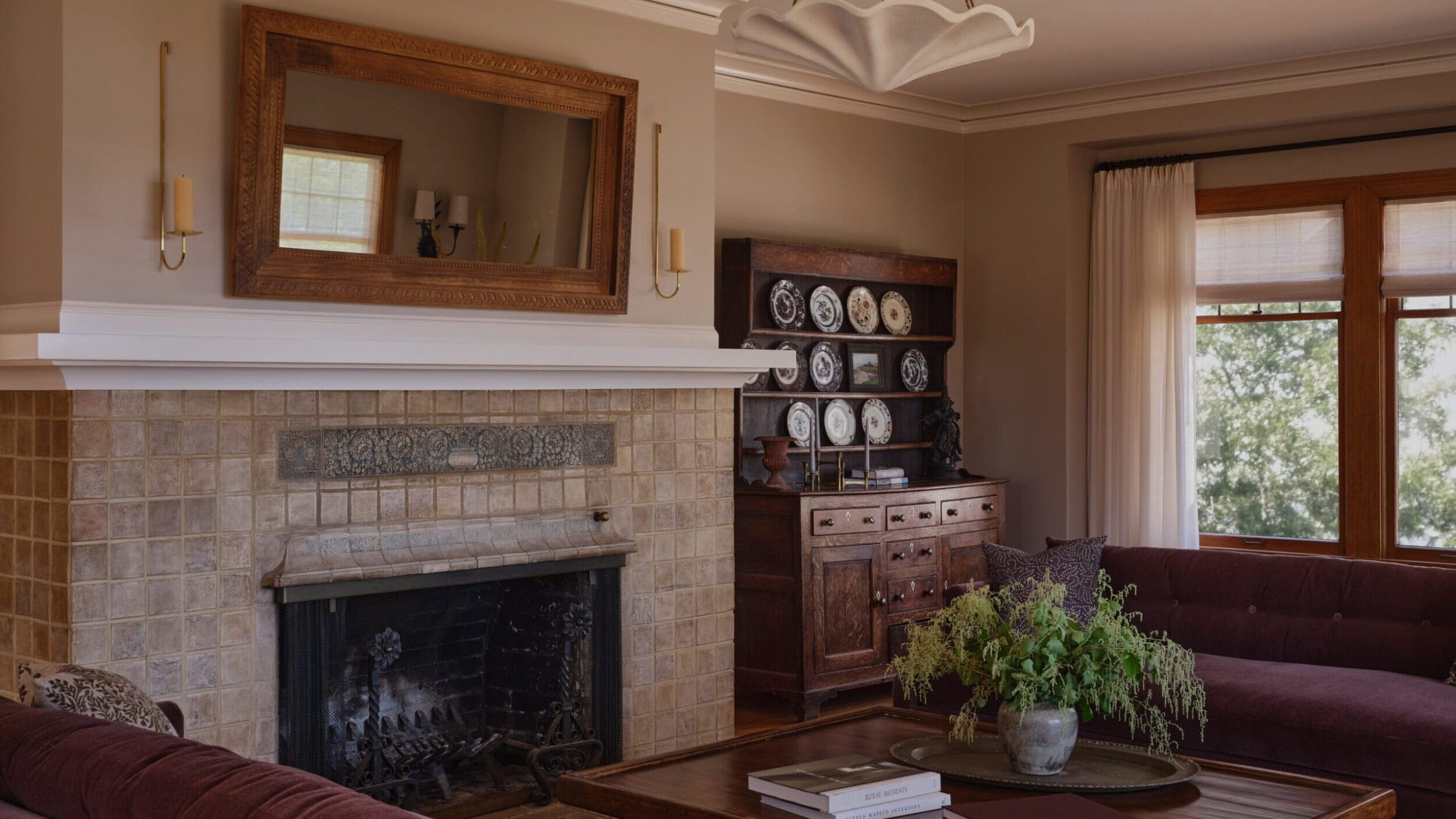 A cozy living room with a fireplace, wooden furniture, and decorative plates on a shelf. Large mirror above the fireplace and green plants on the coffee table.