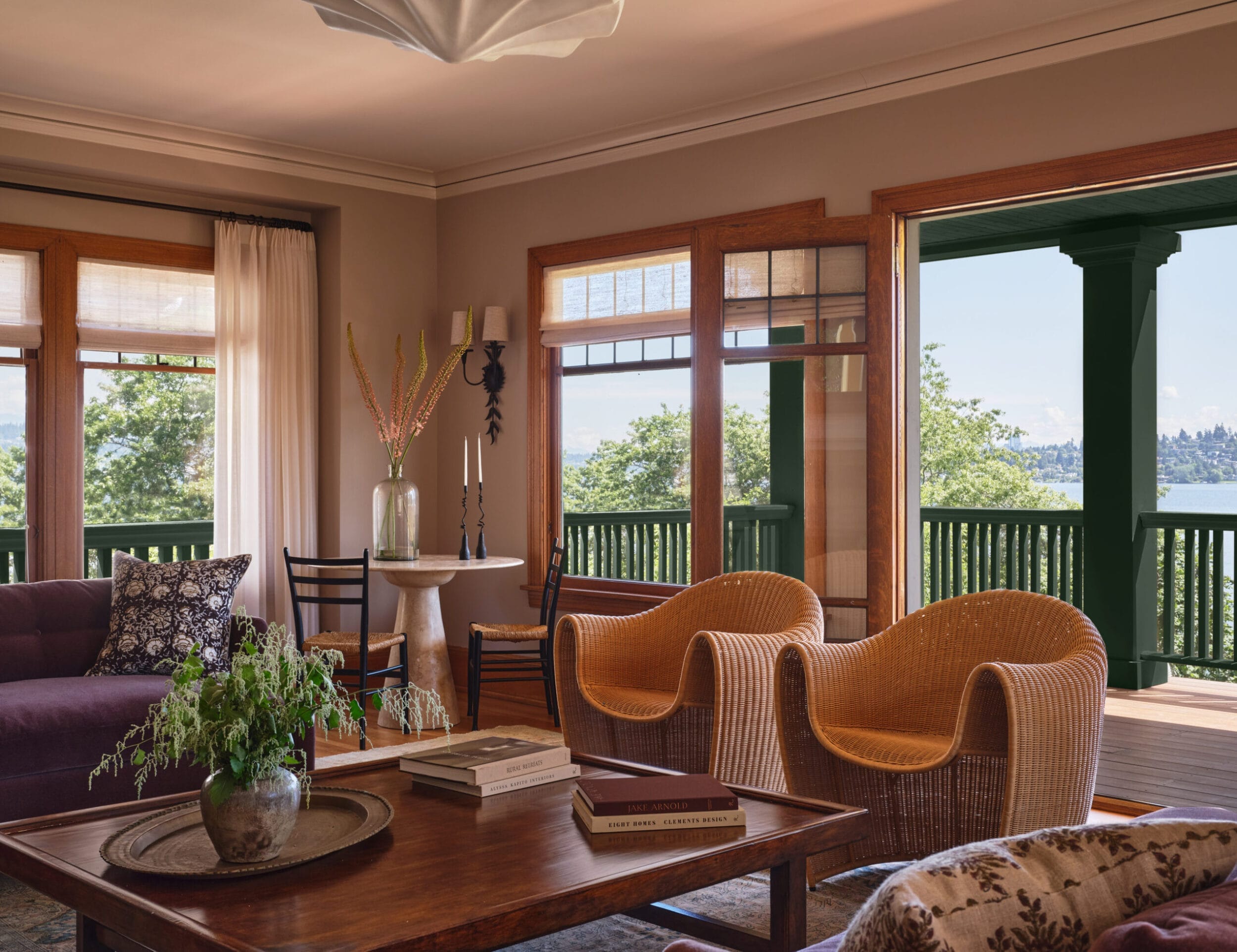 A cozy living room features wicker chairs, a purple sofa, potted plants on a wooden table, and a view of a lake through large windows and an open door to a porch.