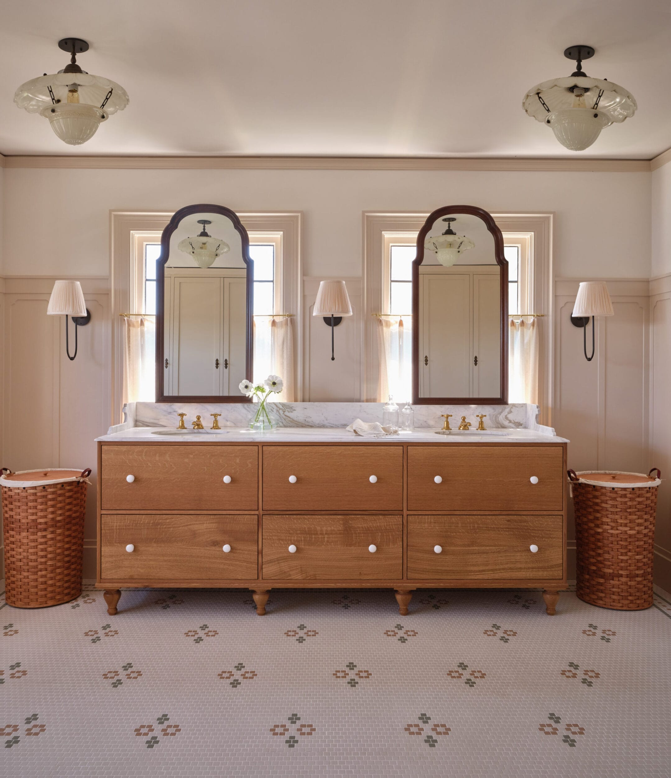 A bathroom with a wooden double vanity, marble countertop, and dual mirrors. There are two wicker laundry baskets and wall sconces, with a patterned tile floor.