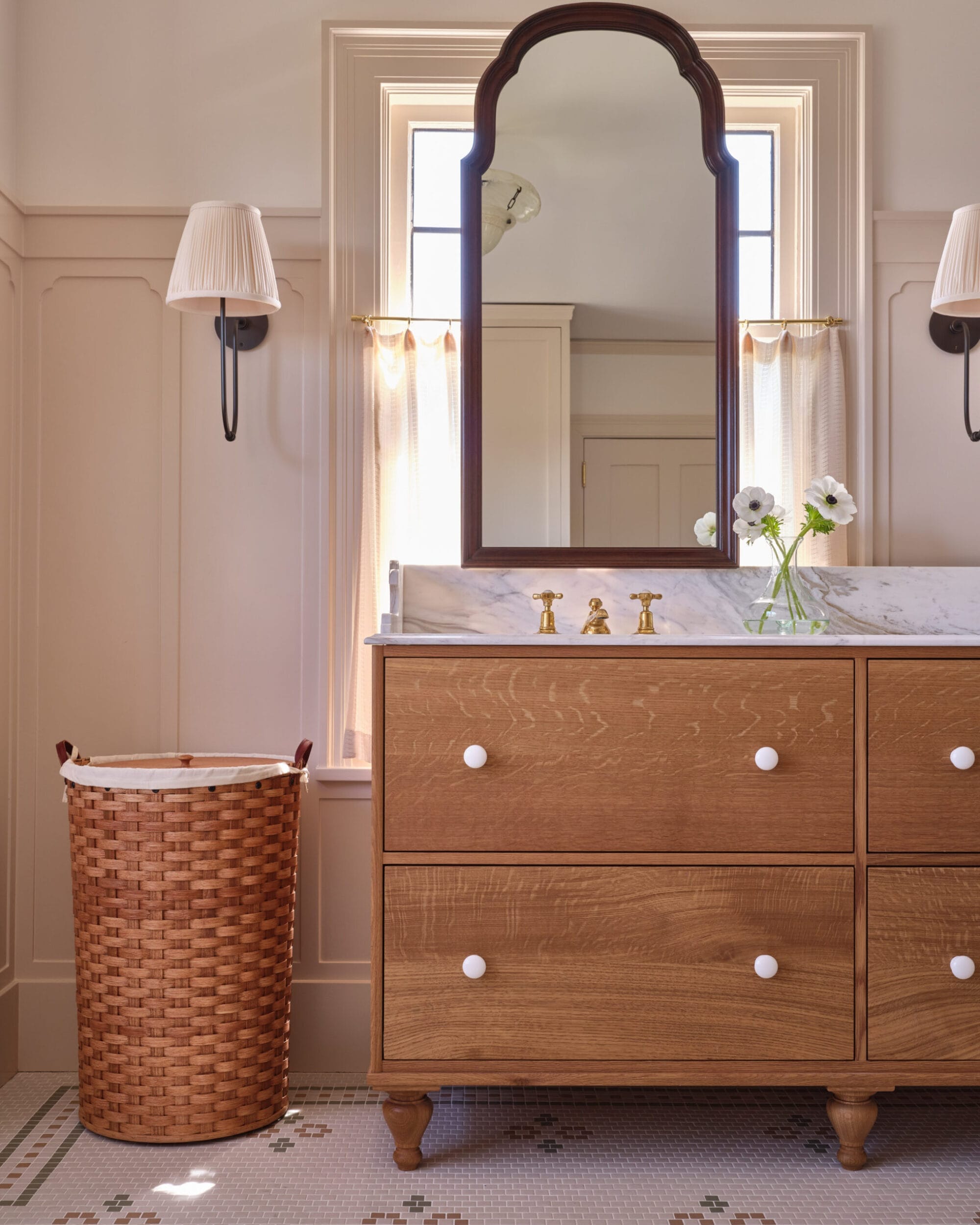 A bathroom with a wooden vanity, marble countertop, arched mirror, and a wicker laundry basket. Two wall lights and a vase with flowers are on the counter.