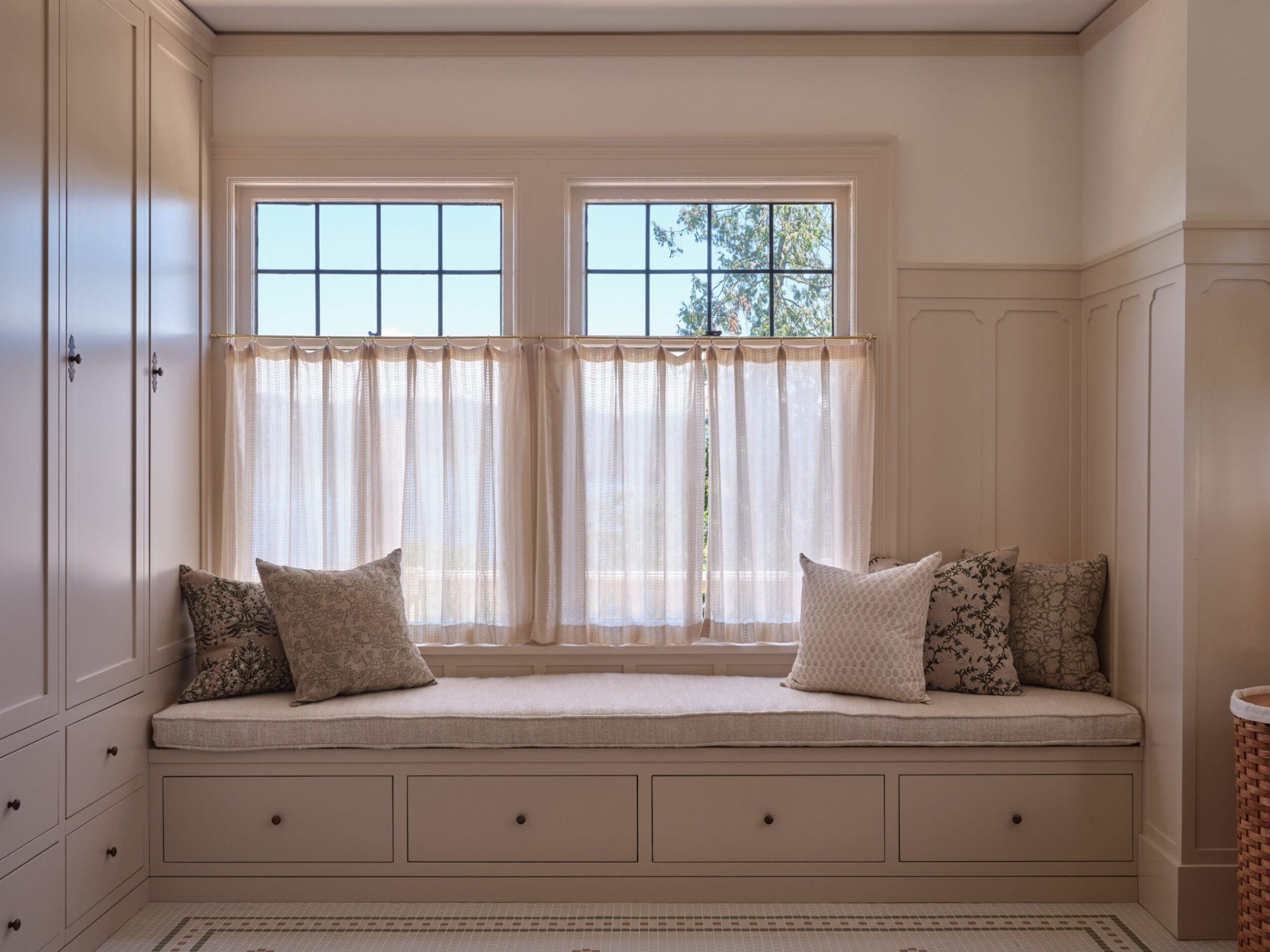 A cozy window nook with beige cushions and pillows, flanked by tall cabinets, under two curtained windows.