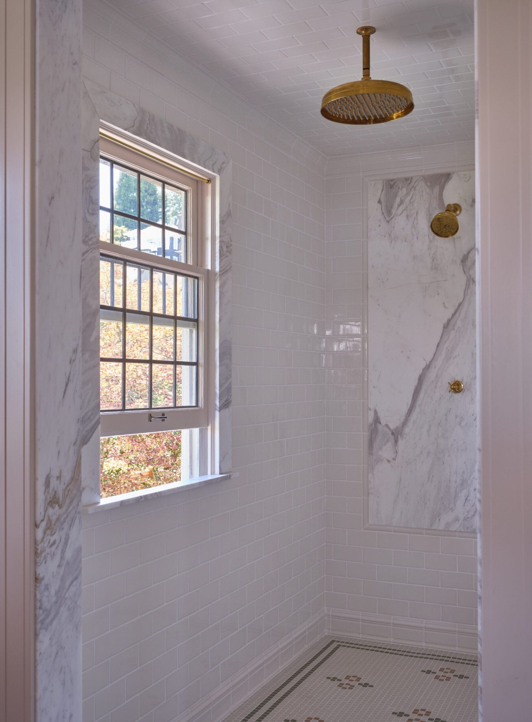 Walk-in shower with marble walls, a square overhead shower head, brass fixtures, and a window with a view of trees.