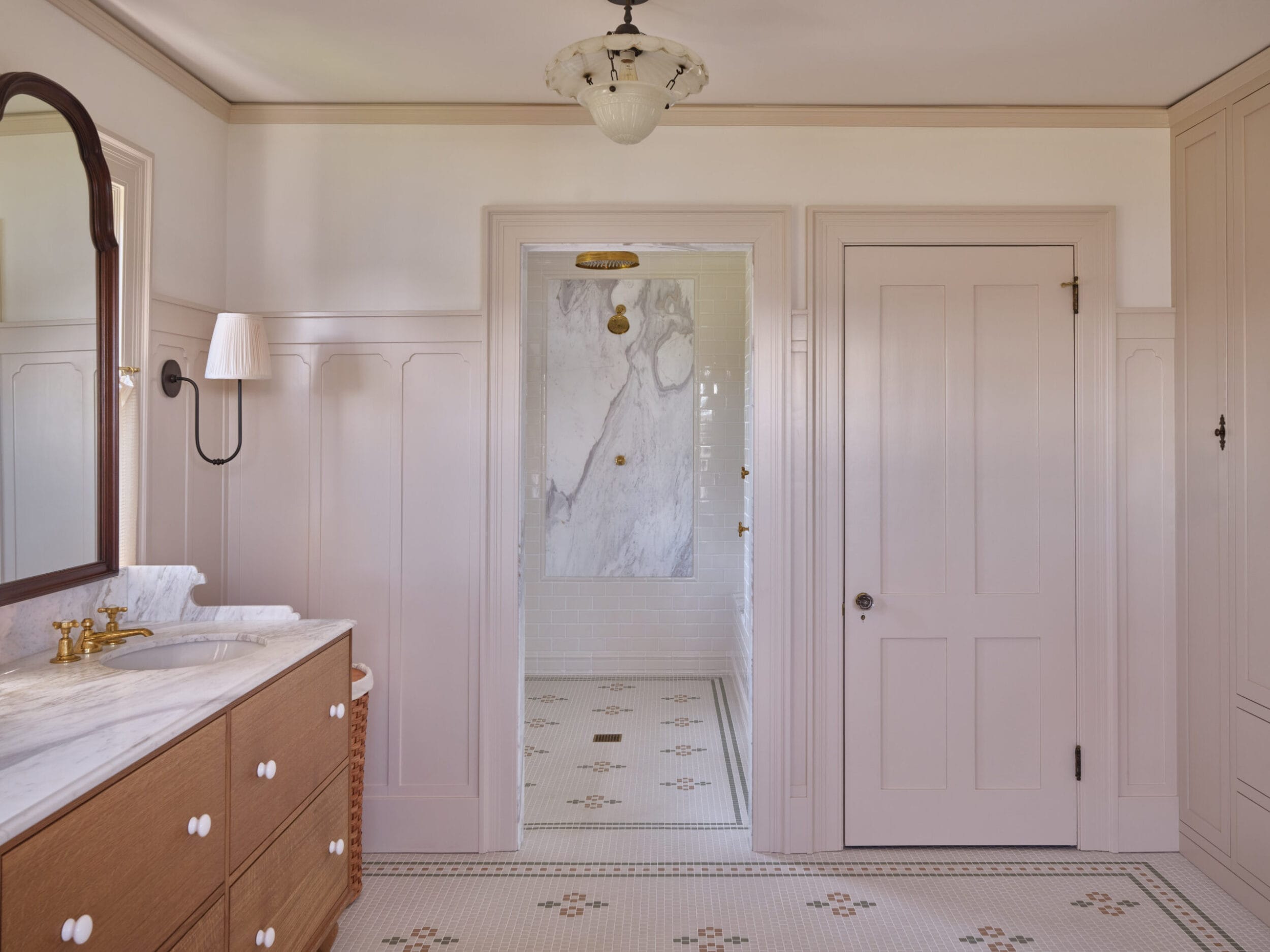 Elegant bathroom with a marble sink, brass faucet, and light fixture. A doorway leads to a marbled shower area with a rainfall showerhead. Neutral tones and patterned floor tiles.