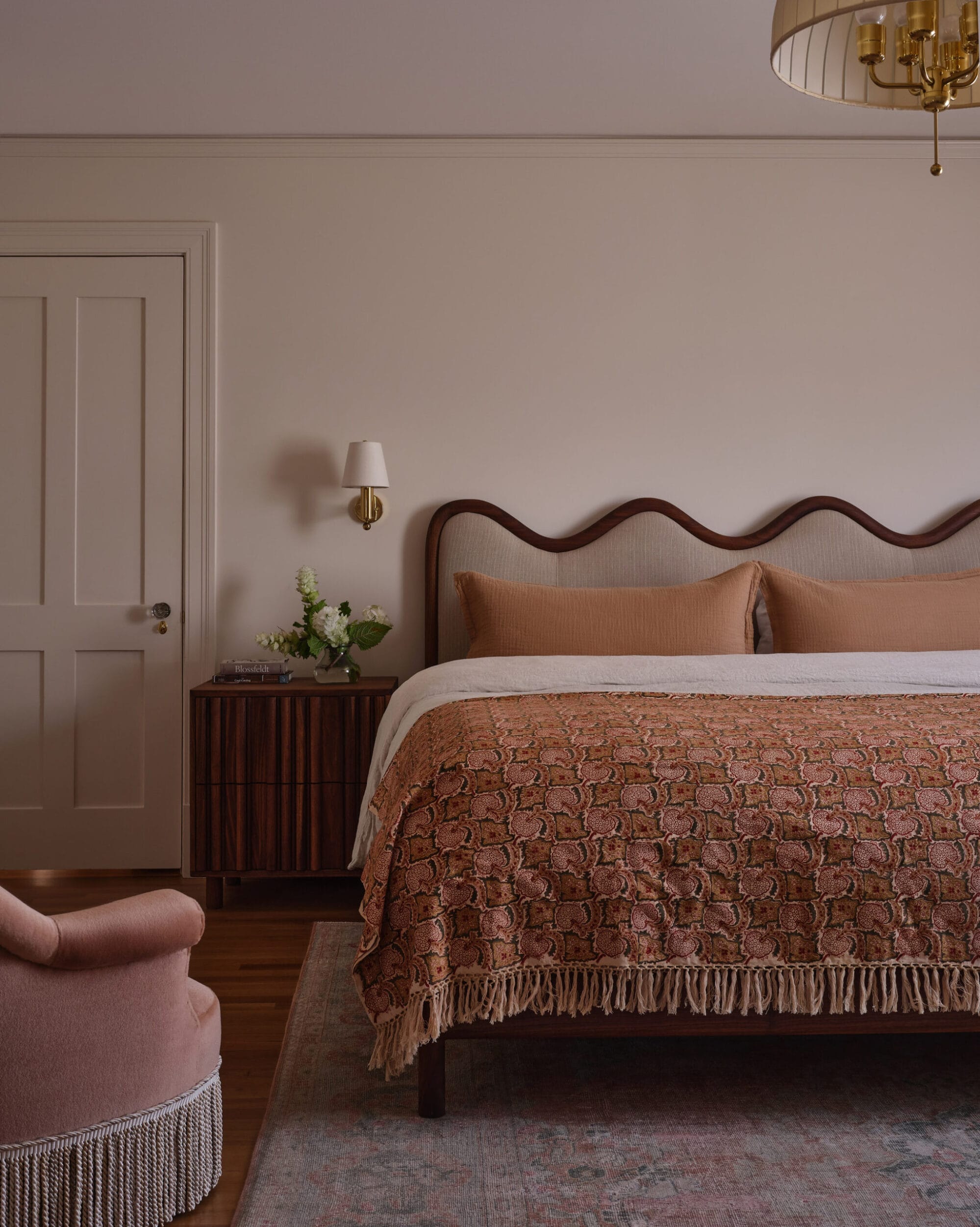 Cozy bedroom with a scalloped headboard, patterned bedspread, and a wooden nightstand with flowers. A chandelier hangs from the ceiling, and a pink armchair is in the foreground.