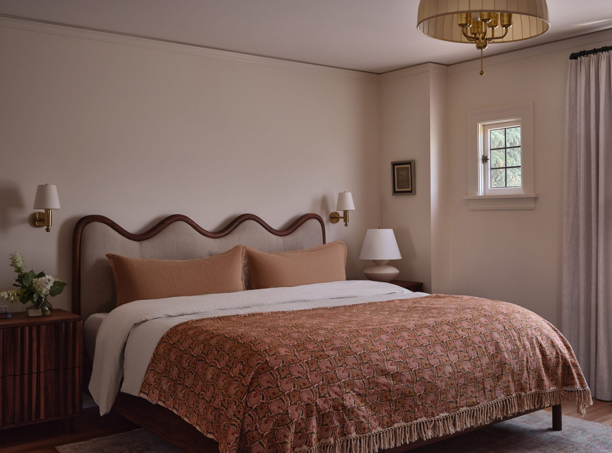 A bedroom with a king-sized bed featuring a patterned blanket and two pillows, flanked by two wooden nightstands with lamps. A window and a ceiling light fixture are also visible.