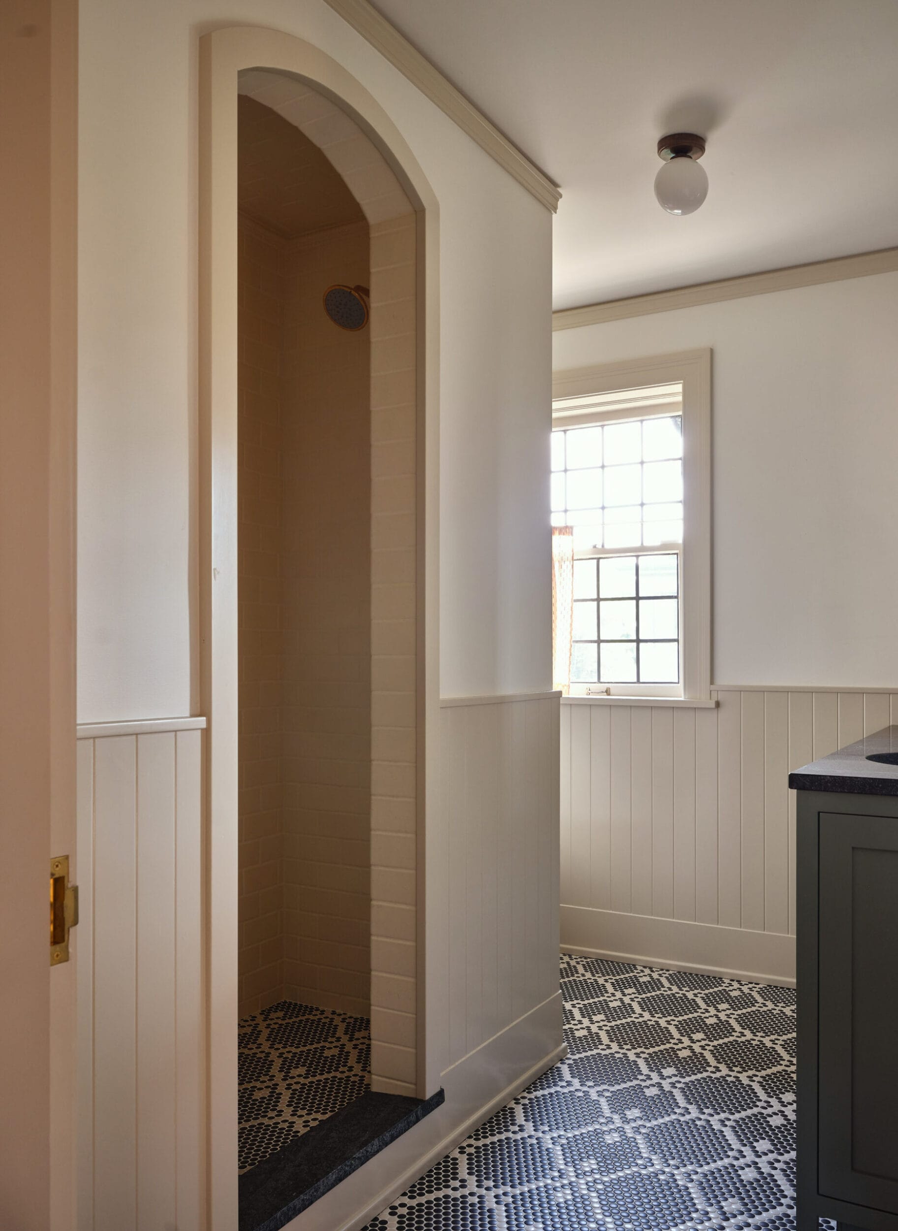 A small alcove shower with an arched entrance, surrounded by light beige walls. The room has a window, a bulb fixture, and a patterned black-and-white tile floor.