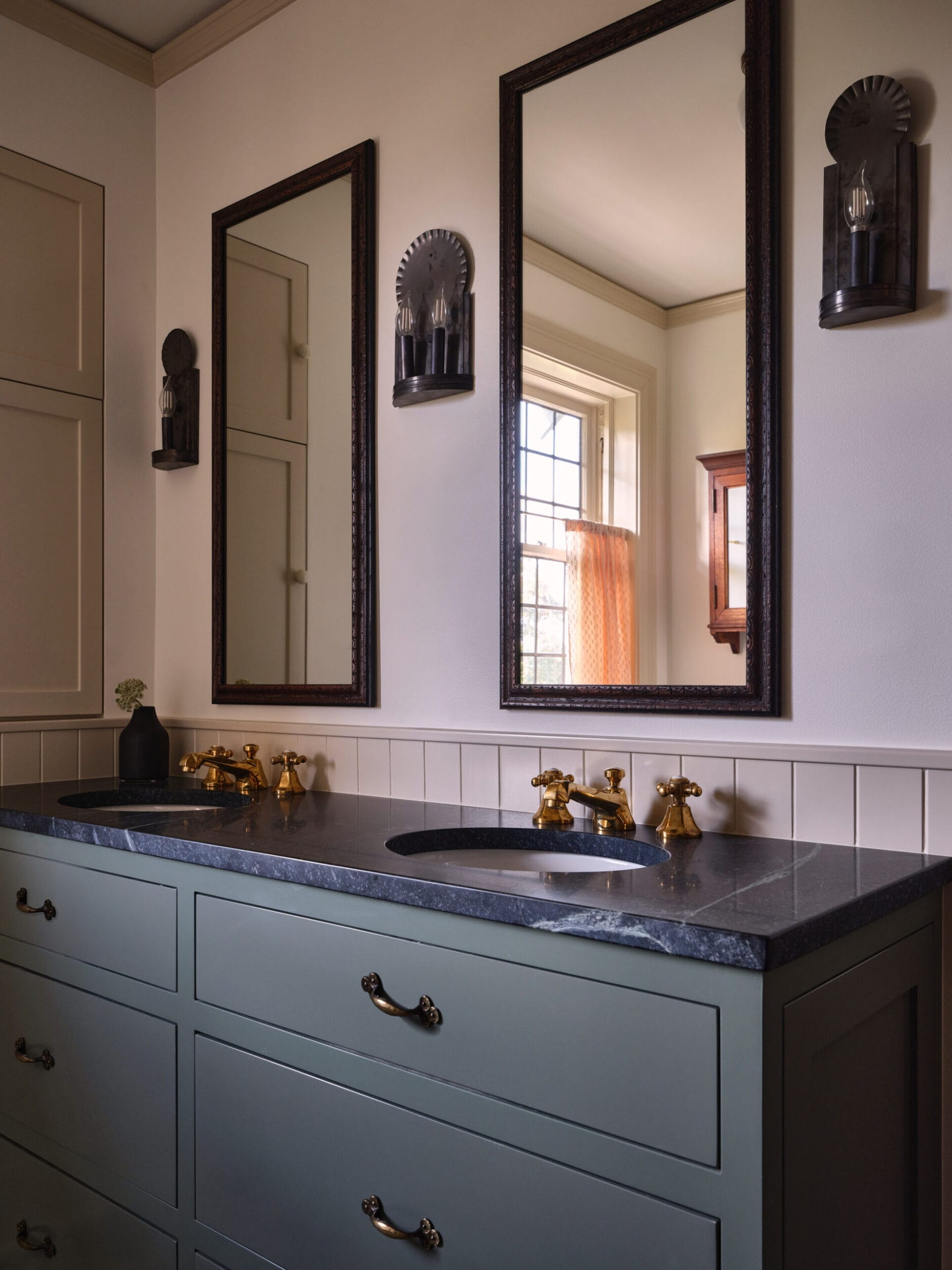 A bathroom with a double sink vanity, brass faucets, two mirrors, and wall sconces. A window with a red curtain is reflected in the mirrors.