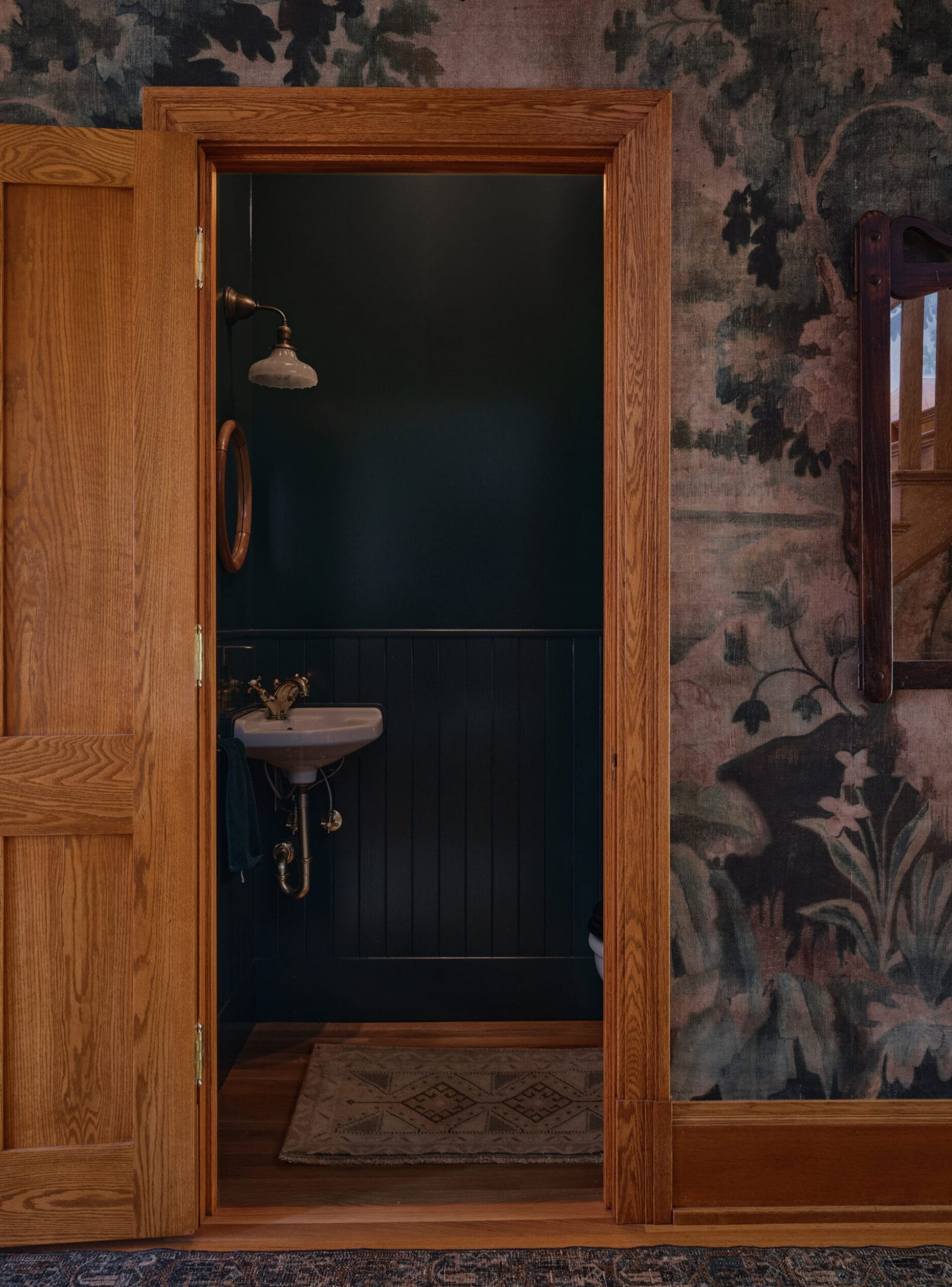 A small, darkly painted bathroom with a white sink and vintage-style showerhead. Wooden door frame and floral wallpaper on the outside wall.