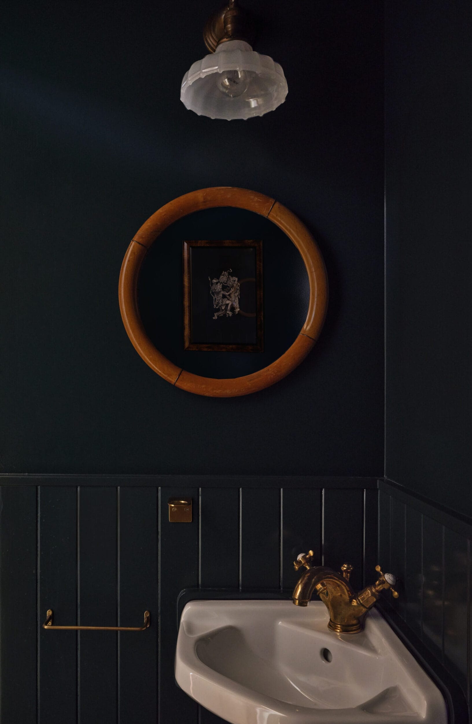 Bathroom with dark walls, a round wooden-framed mirror, white sink, and gold faucet and fixtures. A small decorative picture hangs below a frosted glass light fixture.