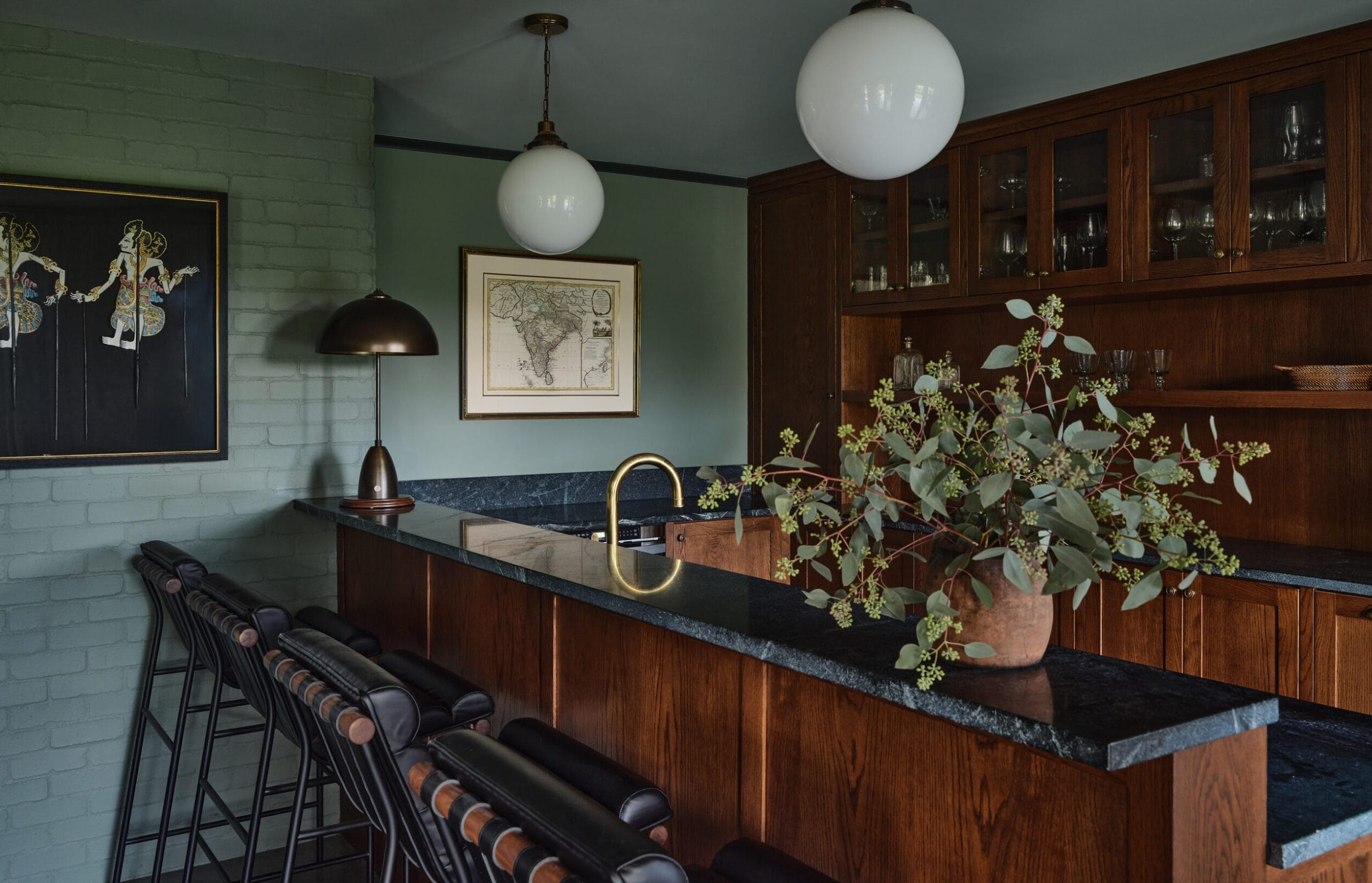 A kitchen with a dark countertop, wooden cabinets, bar stools, and a potted plant. Two round pendant lights hang from the ceiling. Wall art and a lamp are visible in the background.