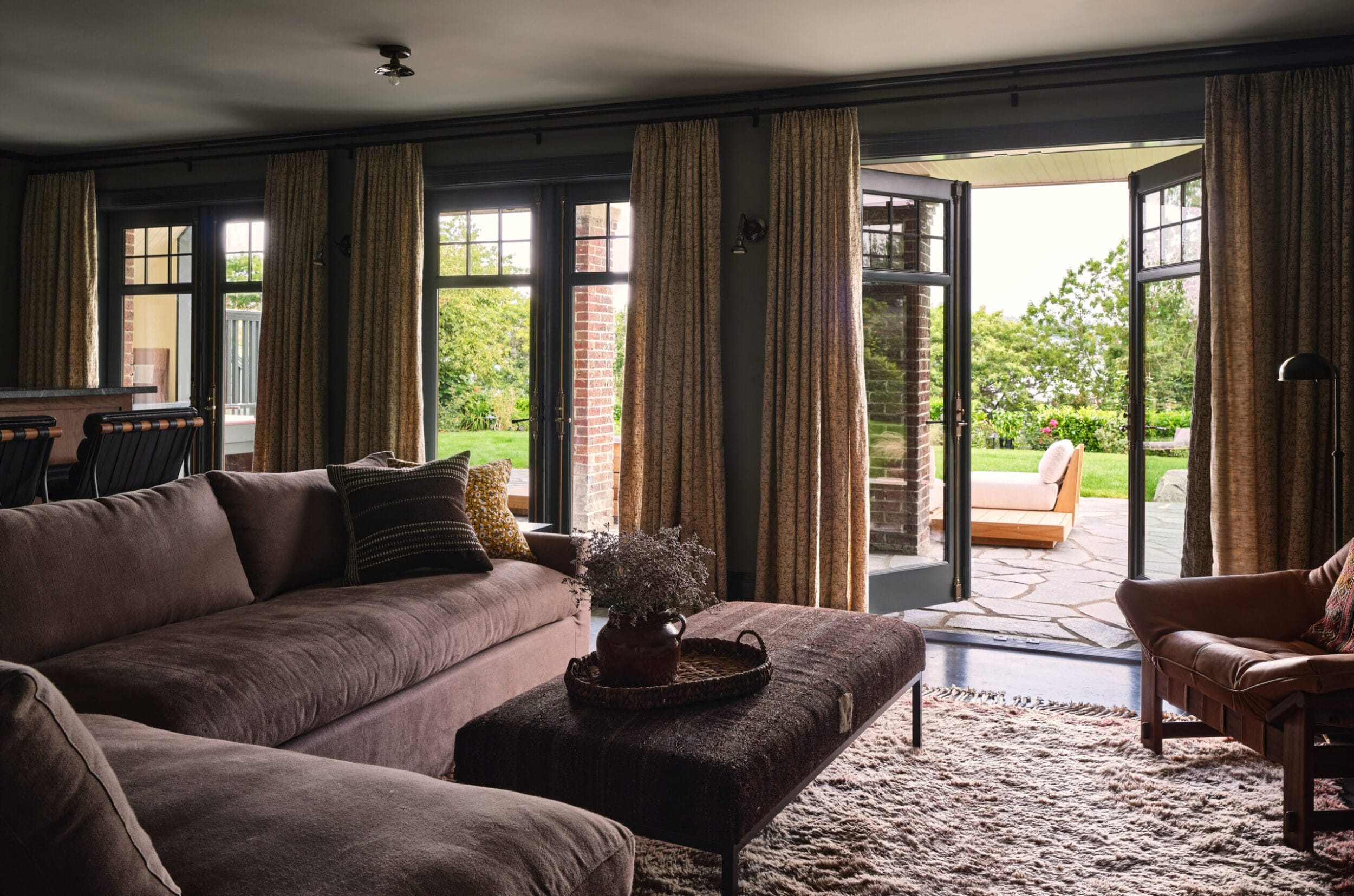 Living room with a large corner sofa, cushioned chairs, and a coffee table. French doors open to a patio with greenery outside. Curtains frame the windows and doors.