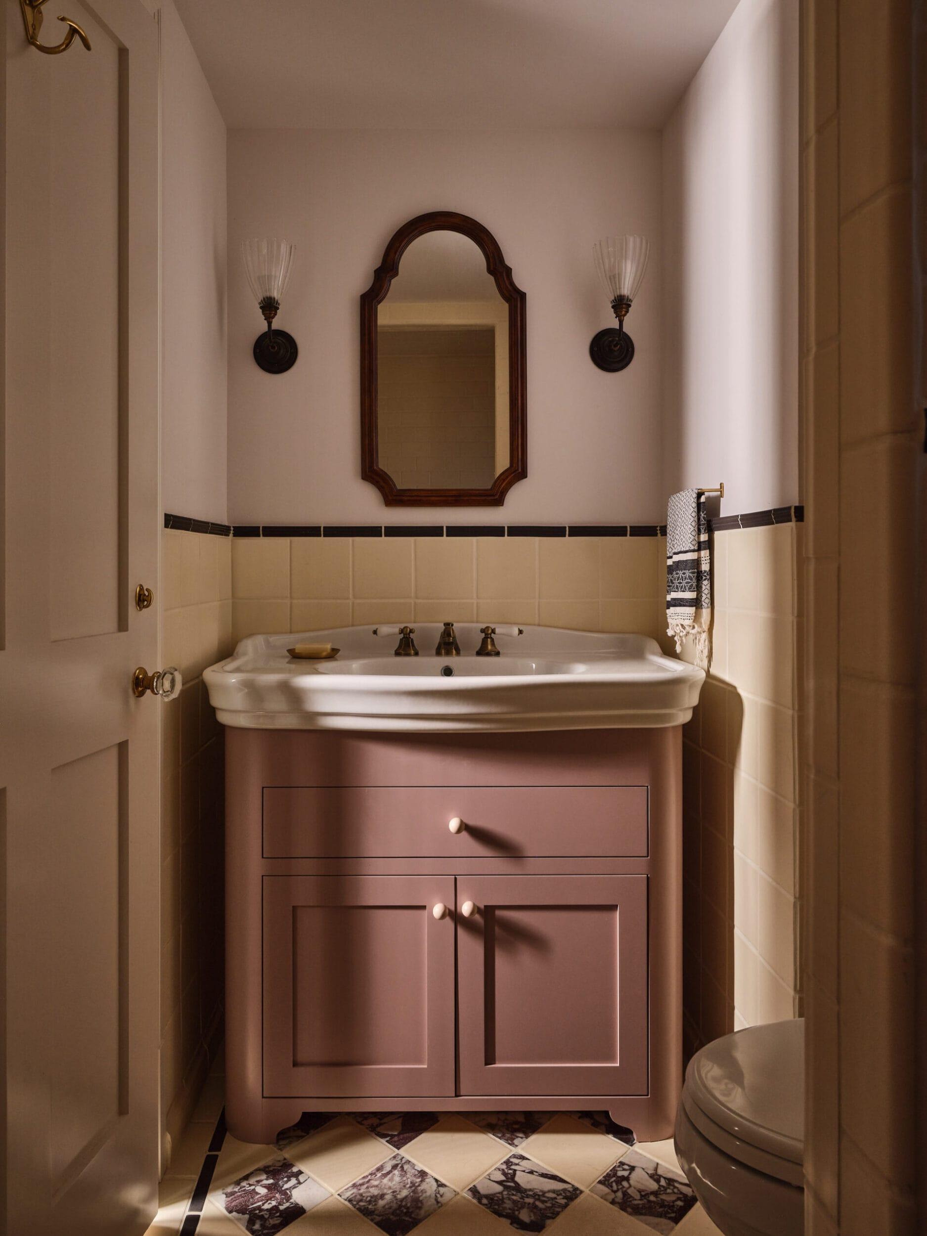 A small bathroom features a vintage sink with a pink vanity, an oval mirror, wall sconces, and black-and-white checkered floor tiles.