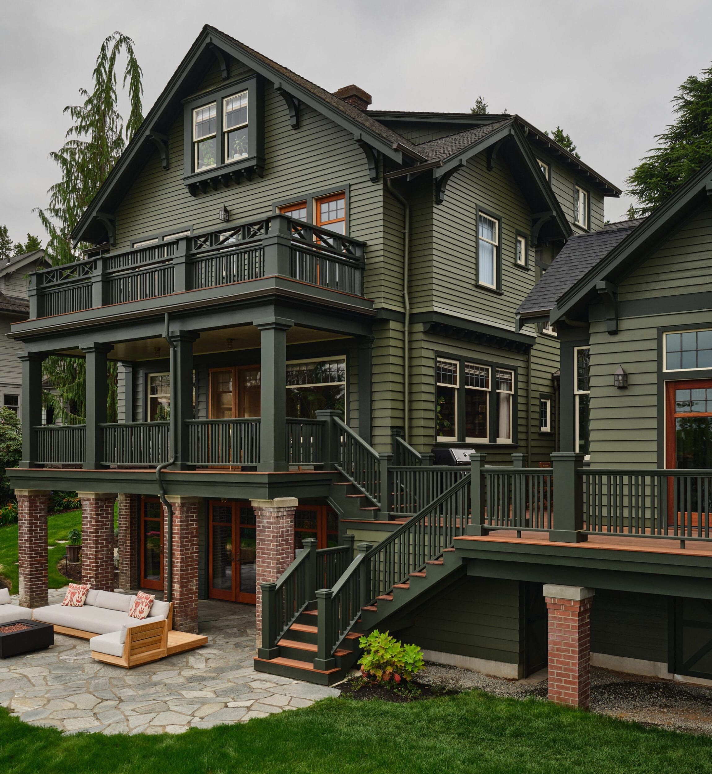 A two-story green house with brick accents, a large balcony, and wooden stairs leading to a paved patio with outdoor seating on a grassy lawn.