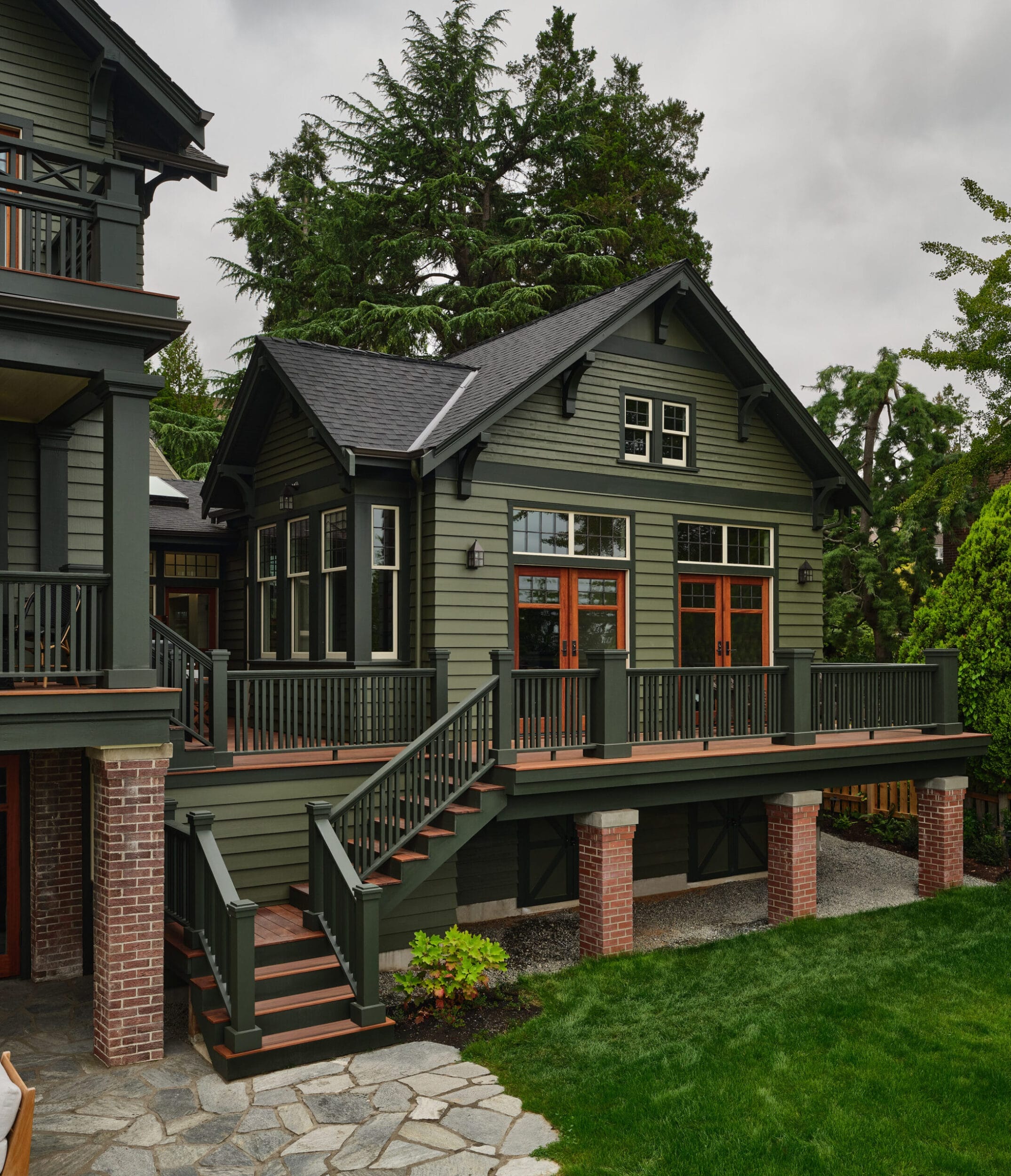 A green two-story house with wooden accents and a large deck, surrounded by trees and a stone path leading to the lawn.