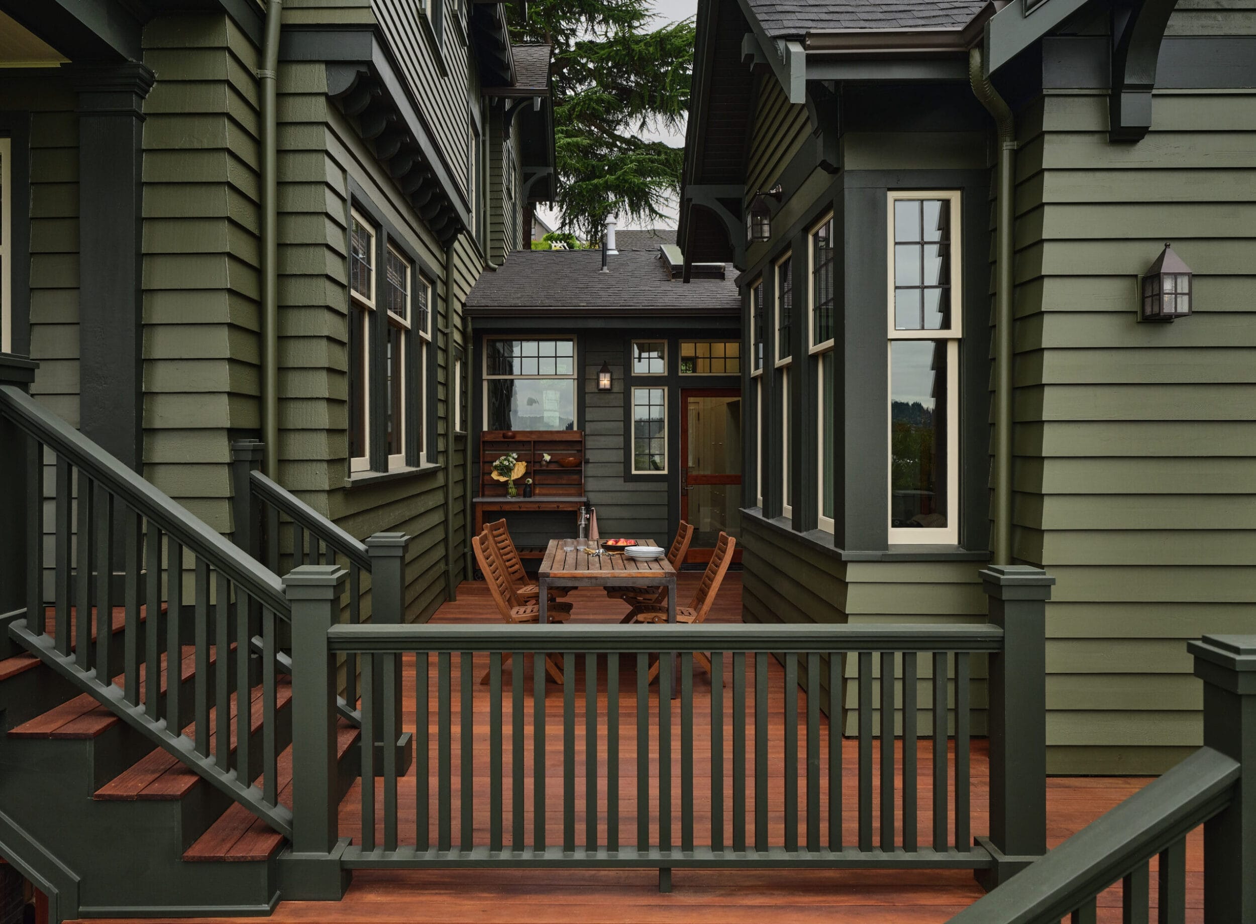 A green wooden house with a porch featuring a wooden dining table and chairs, surrounded by stairs and railings.