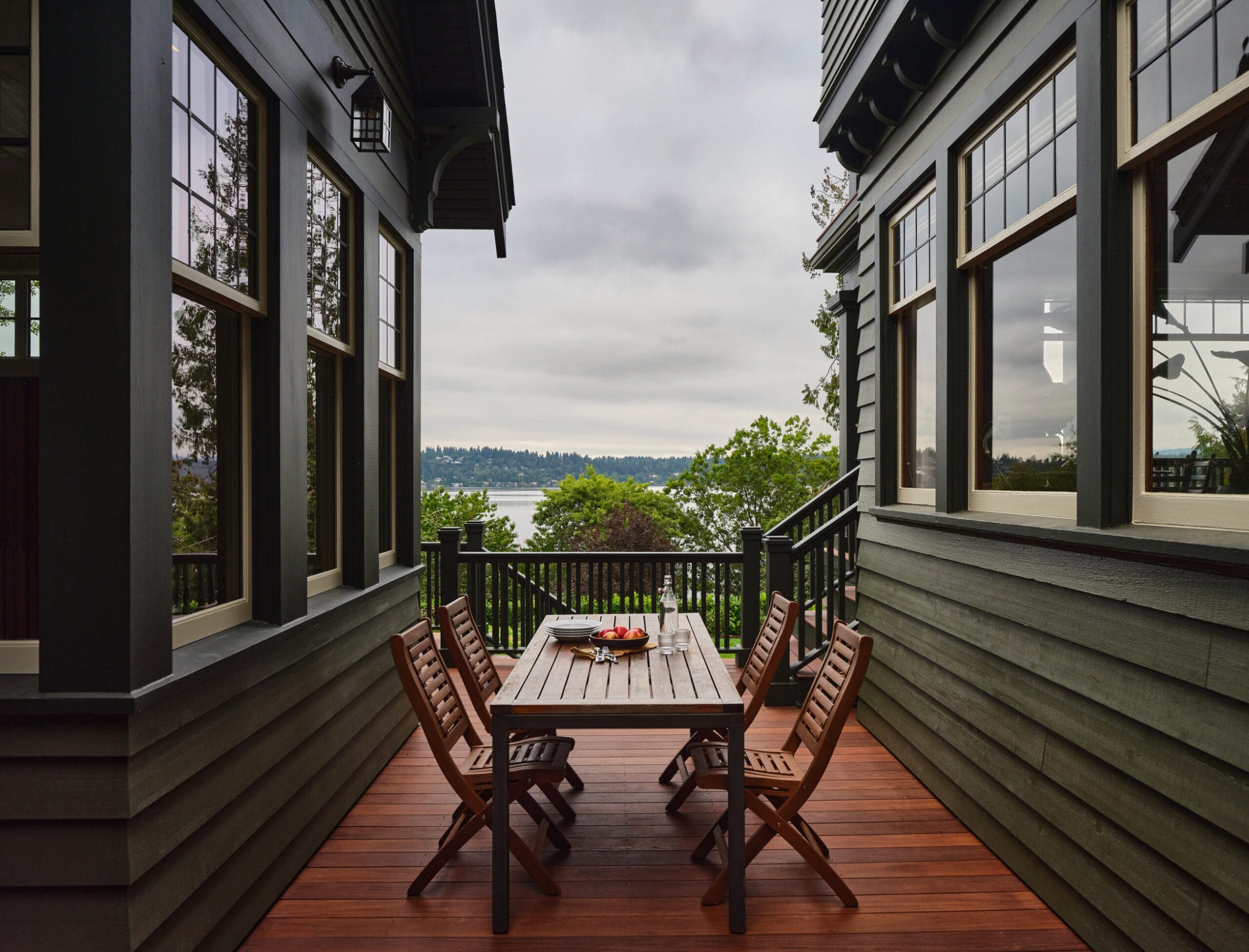 Wooden deck with a dining table and chairs between two dark green buildings, overlooking a view of trees and a body of water in the distance.