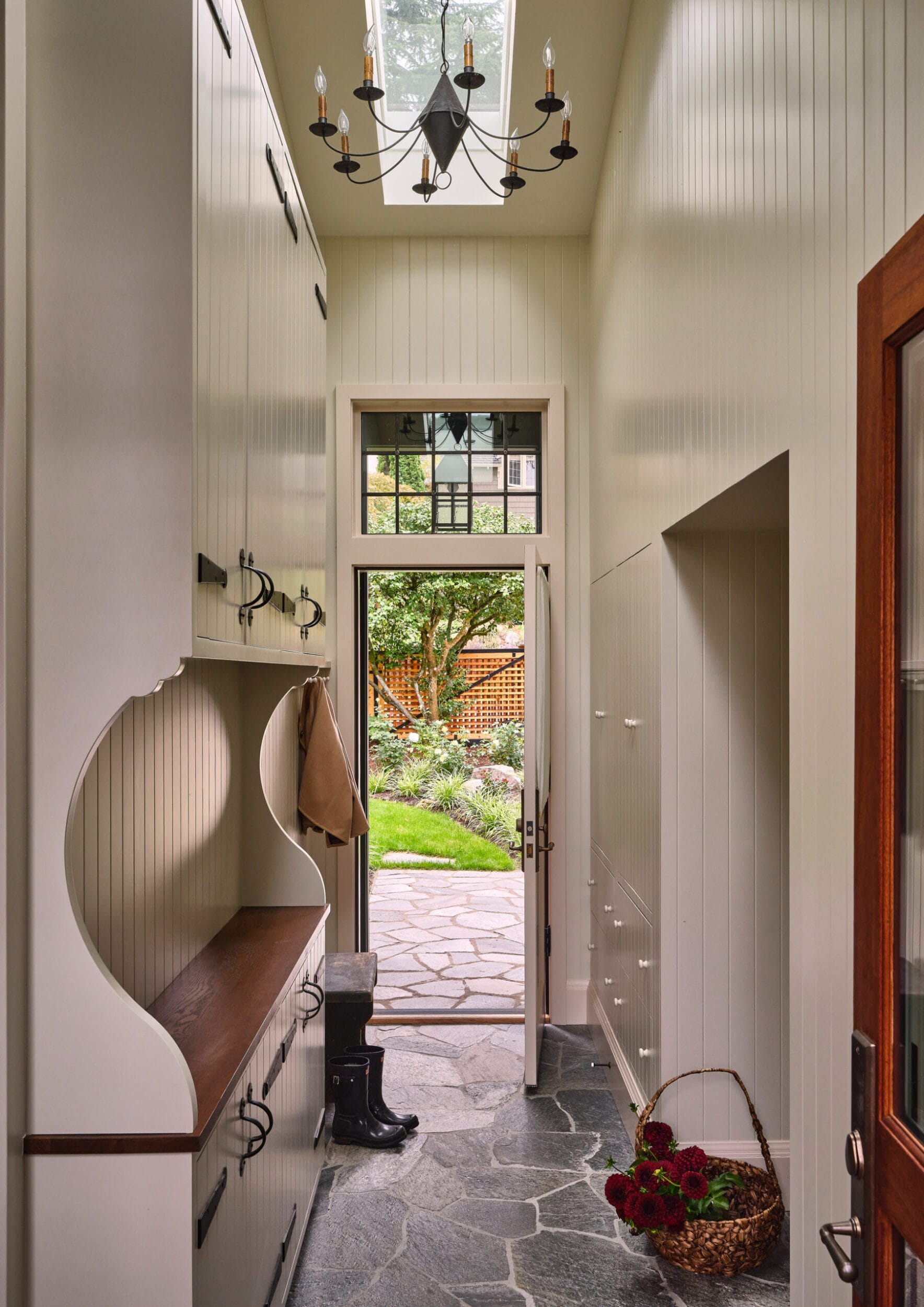 Narrow mudroom with white paneling and wood accents, featuring storage cabinets, a small bench, boots, and a basket. A door leads to a garden with stone paving and greenery.