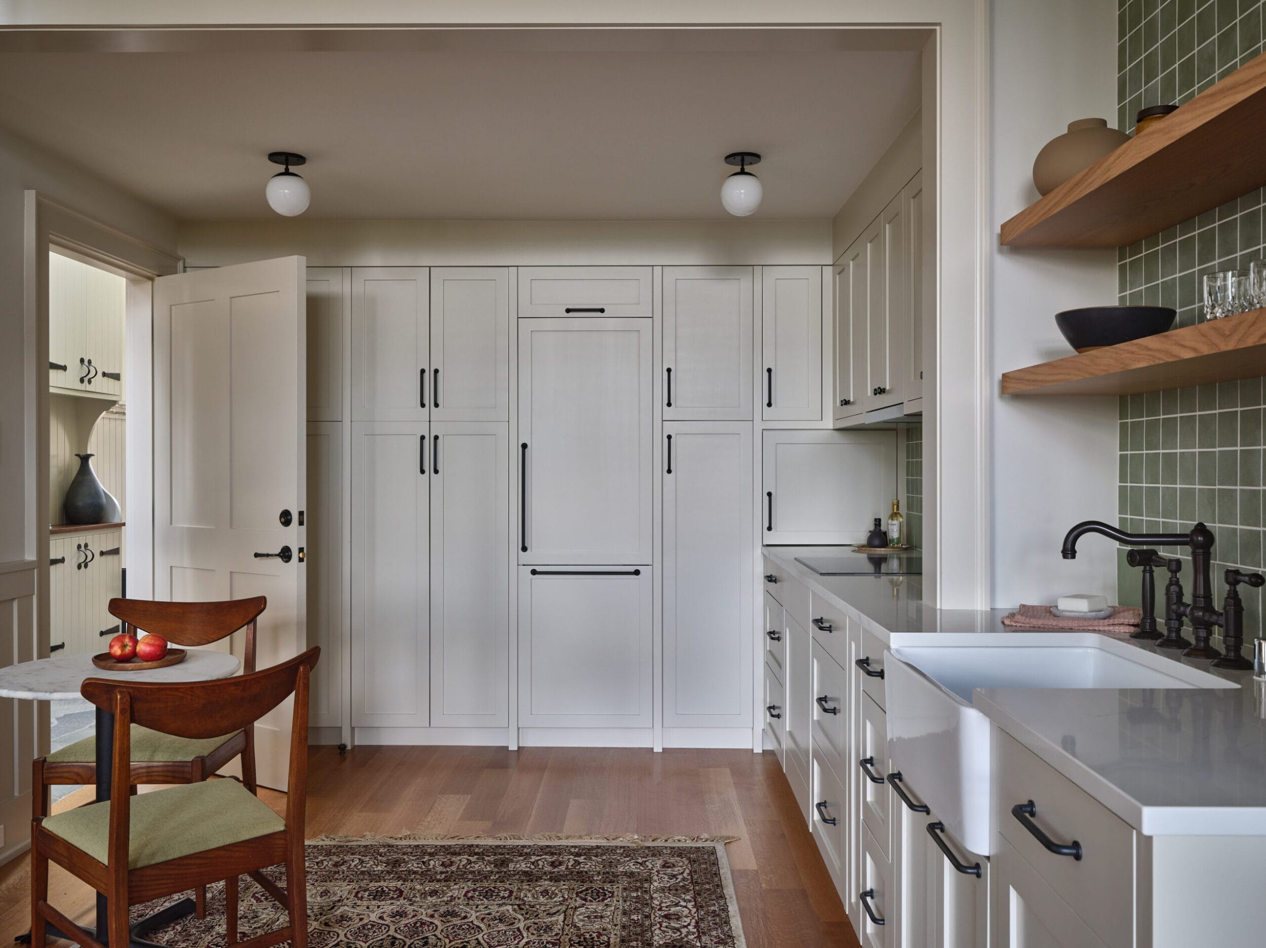 A modern kitchen with white cabinets, green tiled walls, a farmhouse sink, and a small table with chairs. The floor is wood, and the room is well-lit with globe lights.