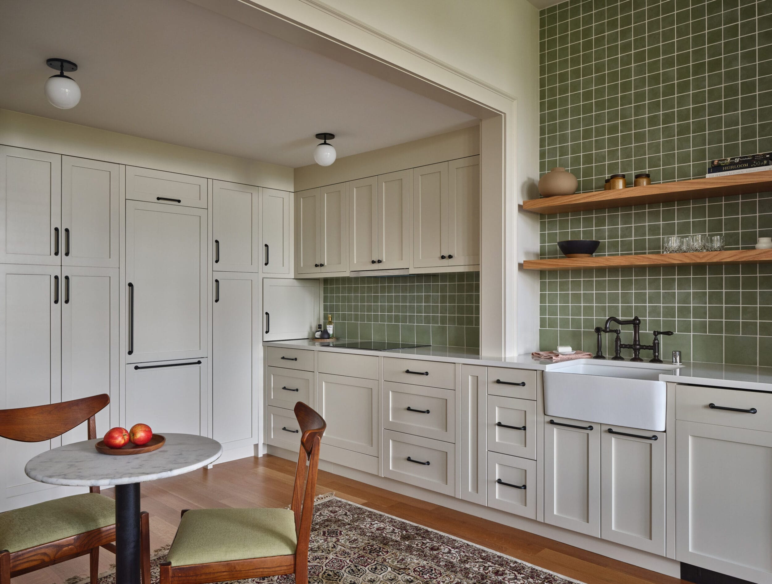 A kitchen with white cabinets, green tiled backsplash, farmhouse sink, and open wooden shelves. A small dining table with chairs and a few apples is placed near the kitchen area.