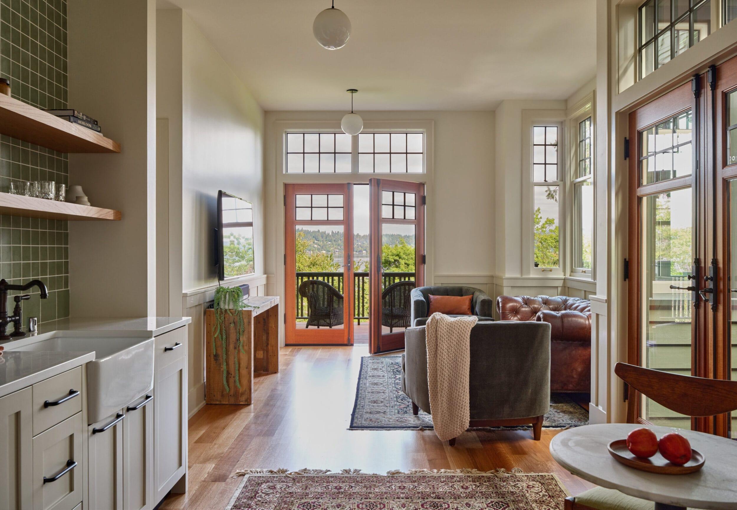 Living room with a sofa, TV, and a small kitchen. Glass doors open to a balcony with a view of trees. Rug on wooden floor and light fixture on the ceiling.