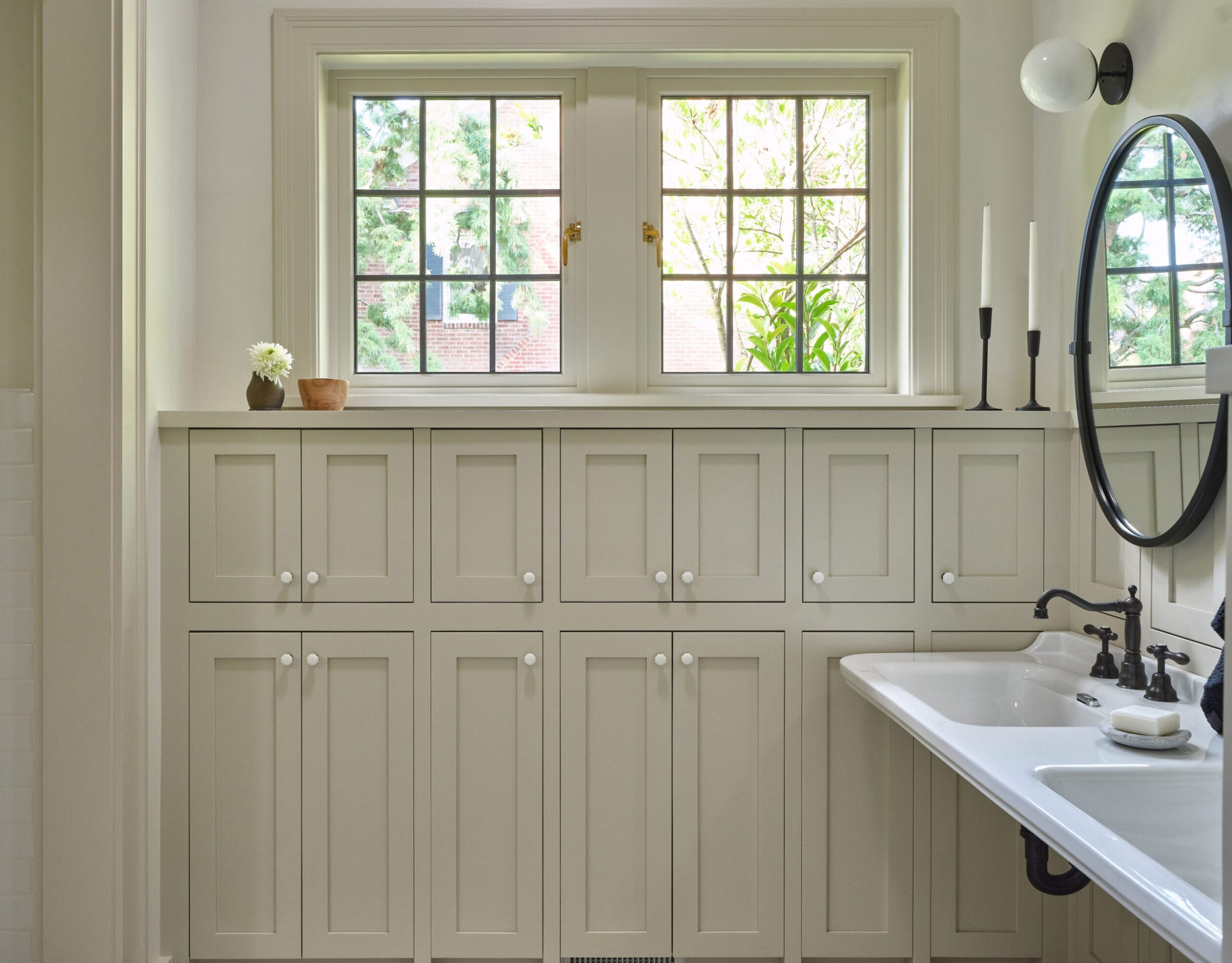 A bathroom with light beige cabinets, a white sink, round mirror, two tall candlesticks, and a small potted plant. There are windows above the cabinets with greenery visible outside.