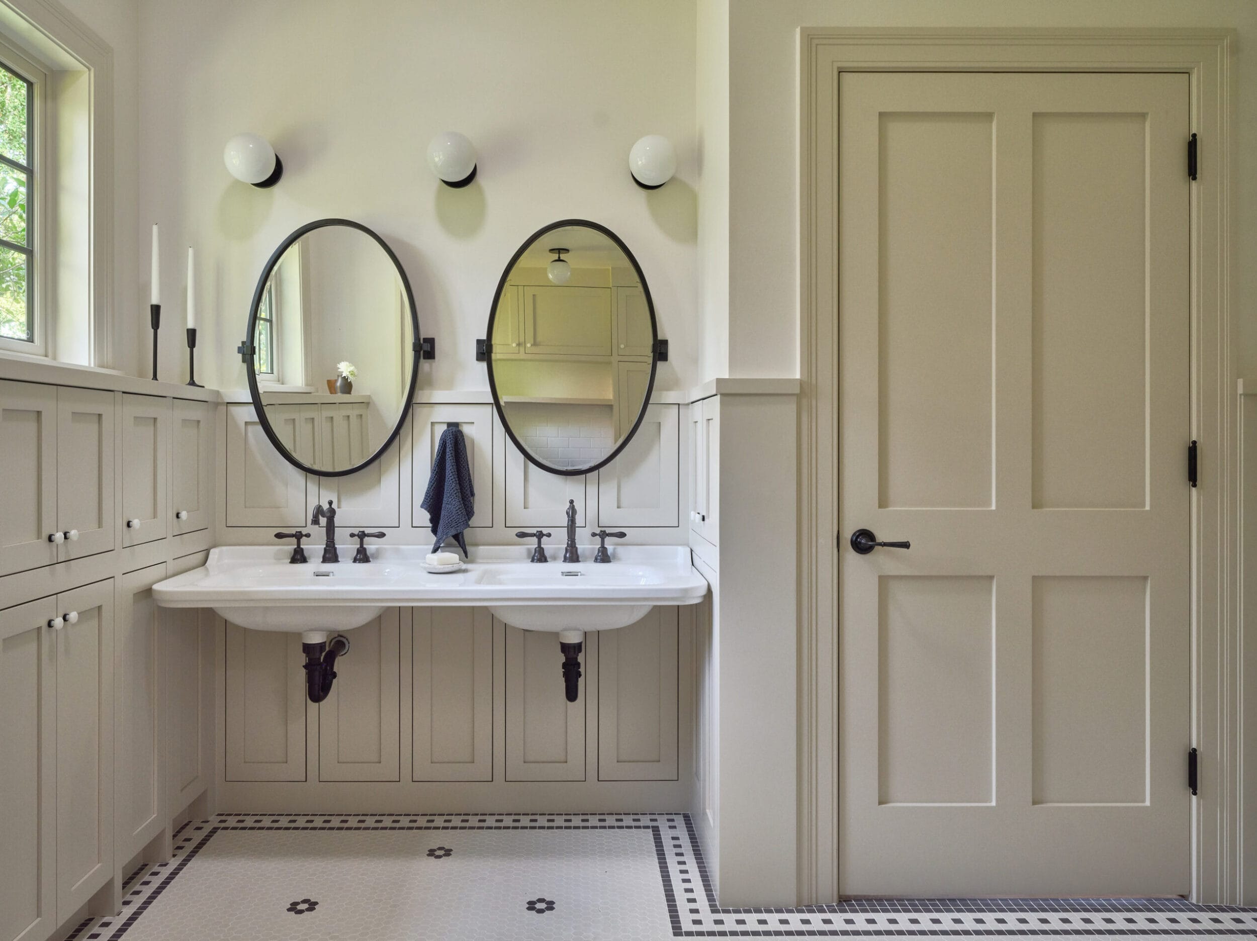 Bathroom with twin sinks, round mirrors, and panelled cabinets. Cream walls and door, with black and white tiled flooring.