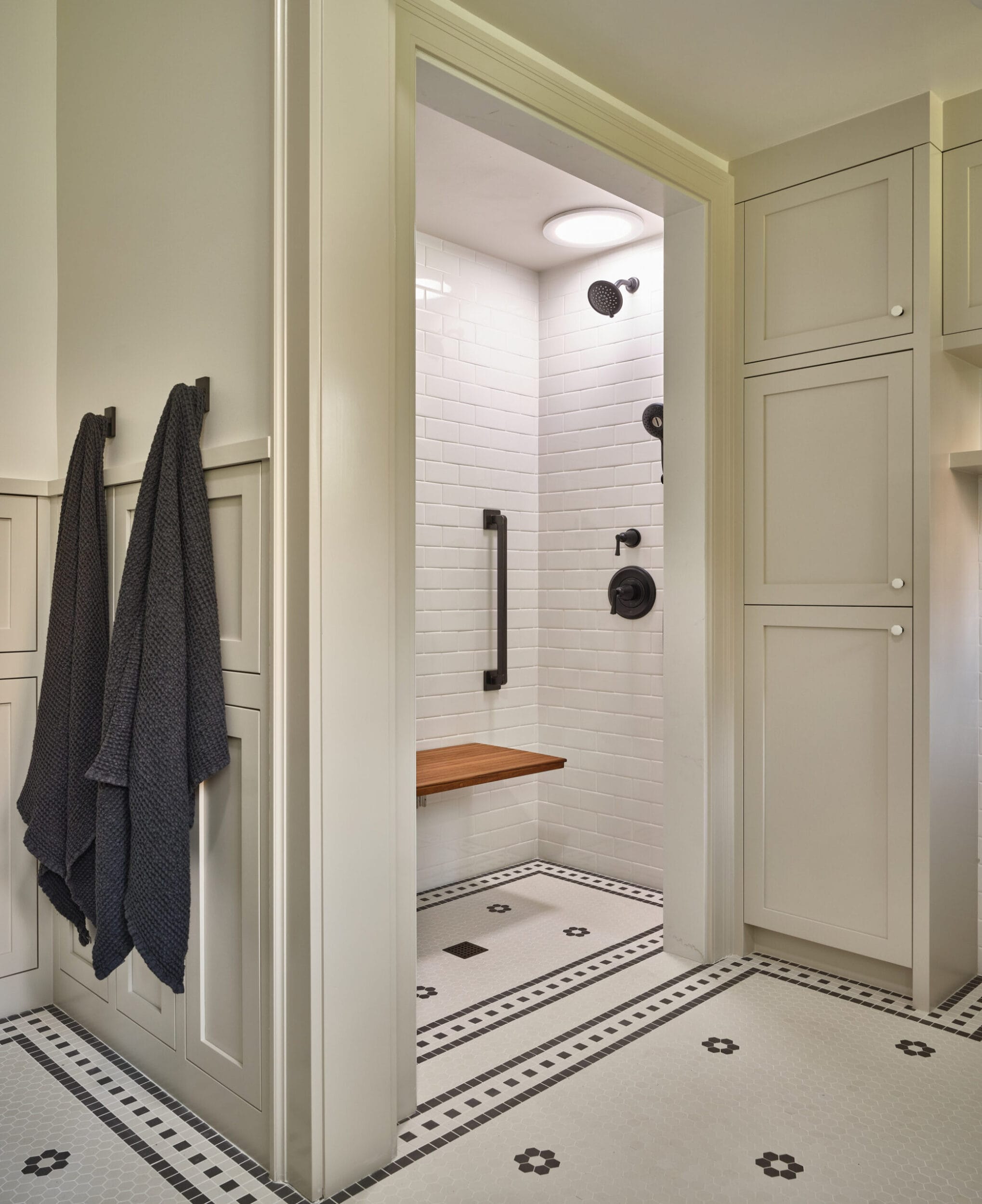 A bathroom features a shower with white subway tiles, a wooden bench, black fixtures, and grab bars. Two navy towels hang outside near built-in cabinetry and a tiled floor with a black floral pattern.