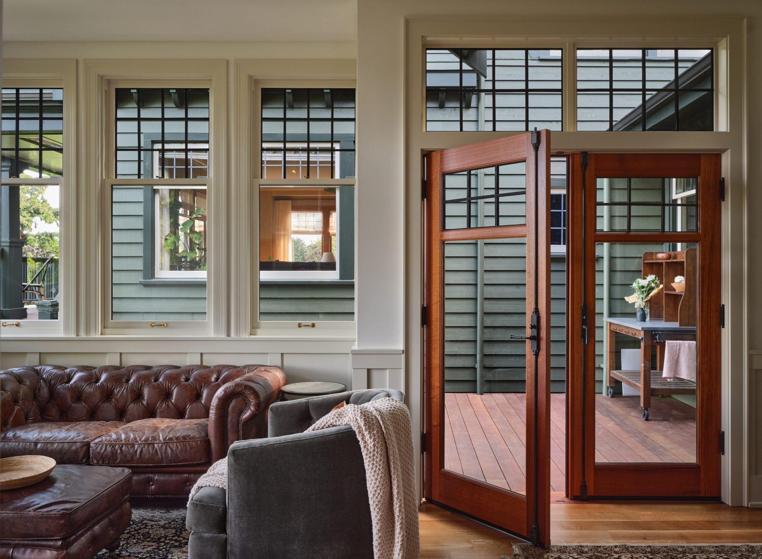 Sunlit living room with brown leather sofas and a view of an outdoor deck through wooden-framed glass doors. Large windows offer natural light.