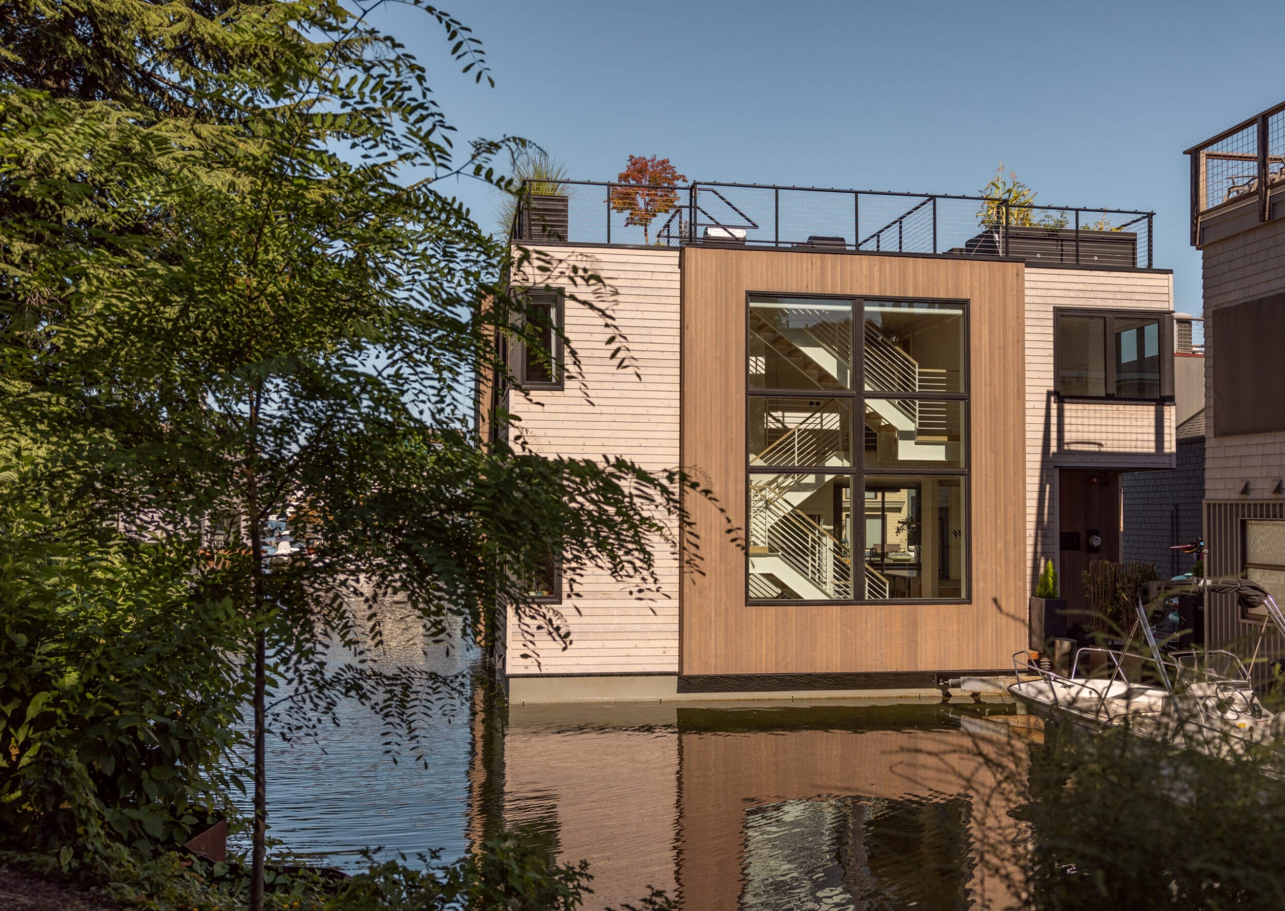 Modern floating house with large windows and rooftop garden, situated on a calm body of water, surrounded by lush greenery.