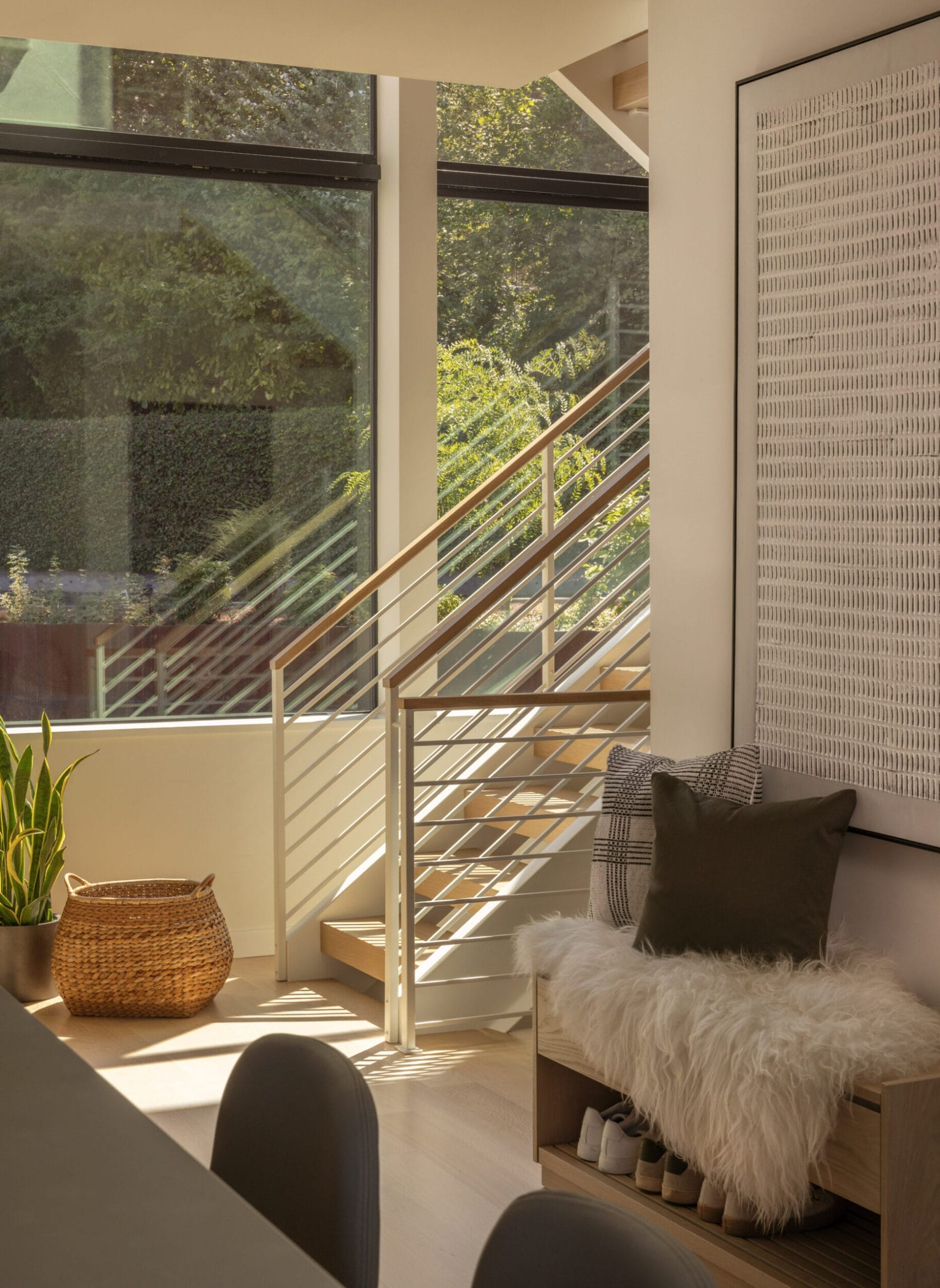 A sunlit interior with a staircase, large windows, a wicker basket, a plant, and a cozy seat with cushions and a throw.