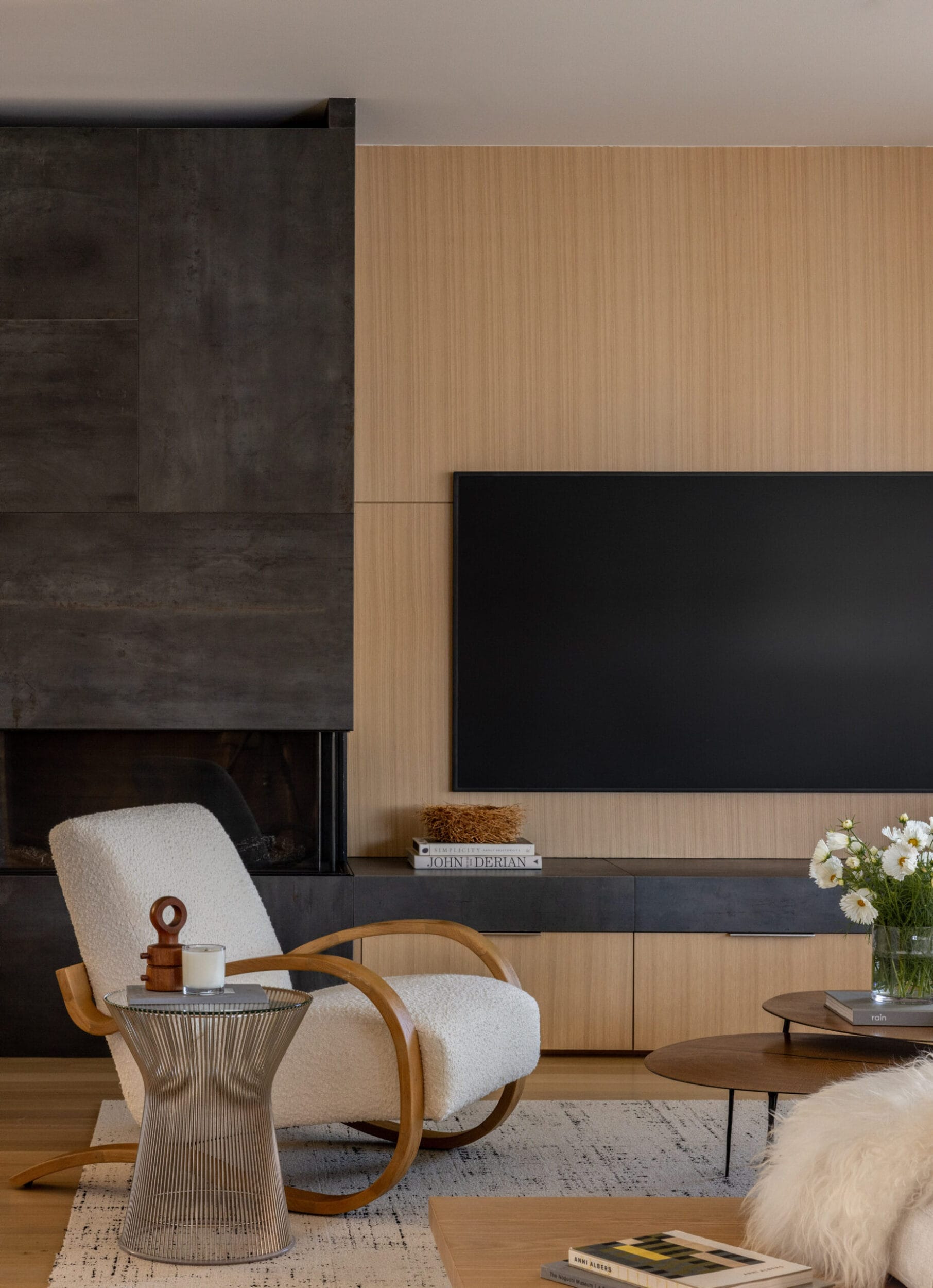 Modern living room with a white armchair, a round side table, a minimalist black fireplace, a large flat-screen TV, and a wooden accent wall. White flowers are on the coffee table.