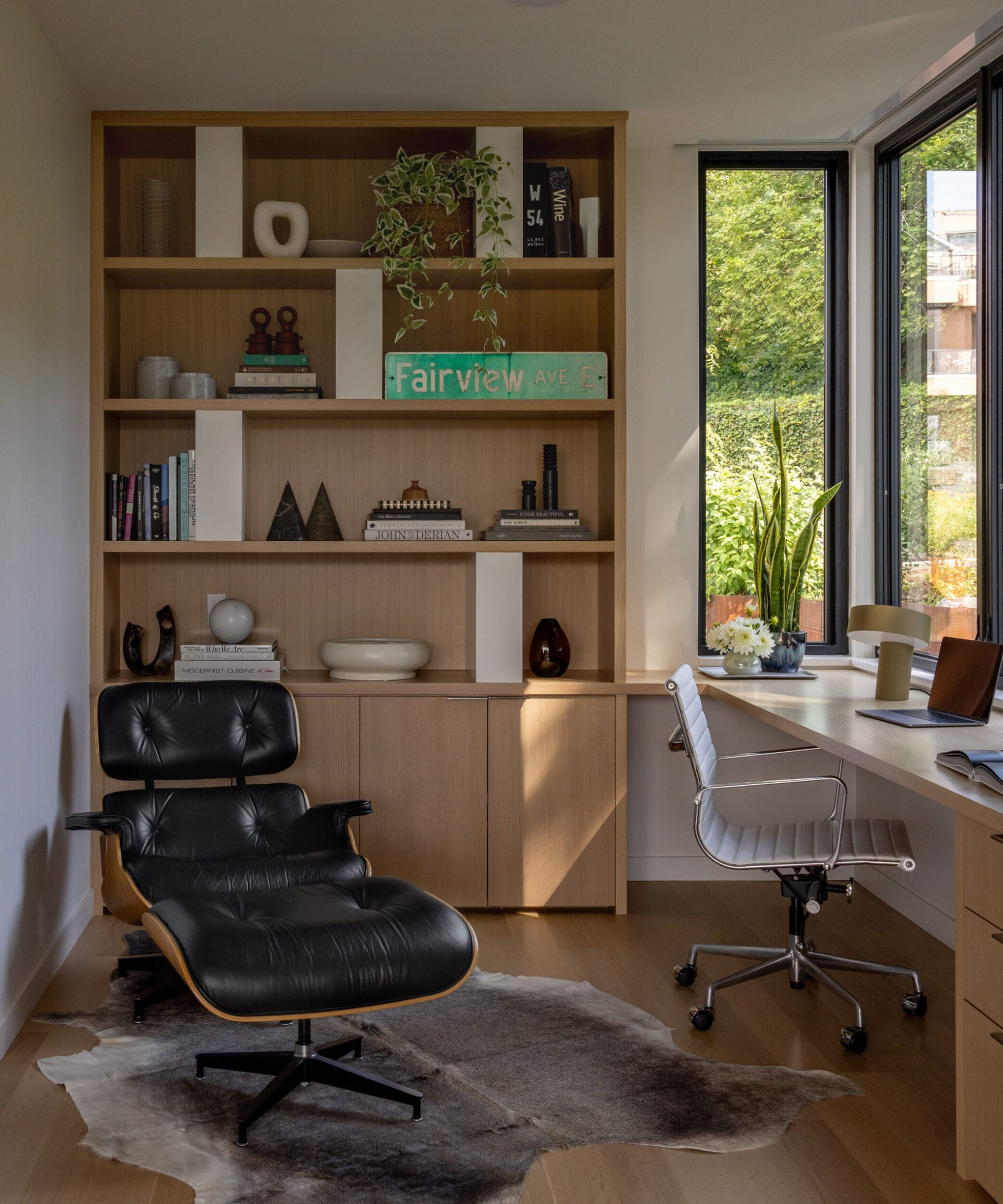 A modern home office with a black lounge chair, a wooden desk and white office chair, bookshelves with decorative items, and large windows overlooking greenery.