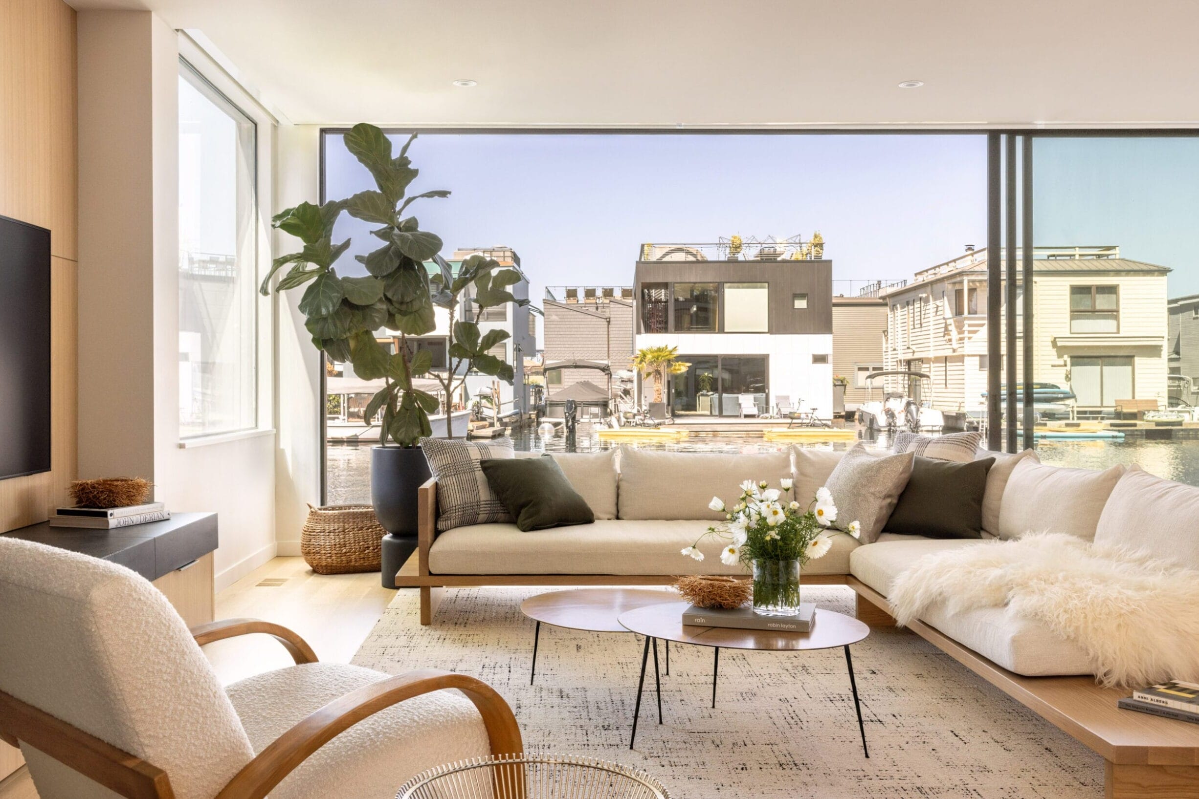 Modern living room with large windows, a beige sofa, a white armchair, a circular coffee table, and a potted plant. Outdoor view of buildings and water is visible through the windows.