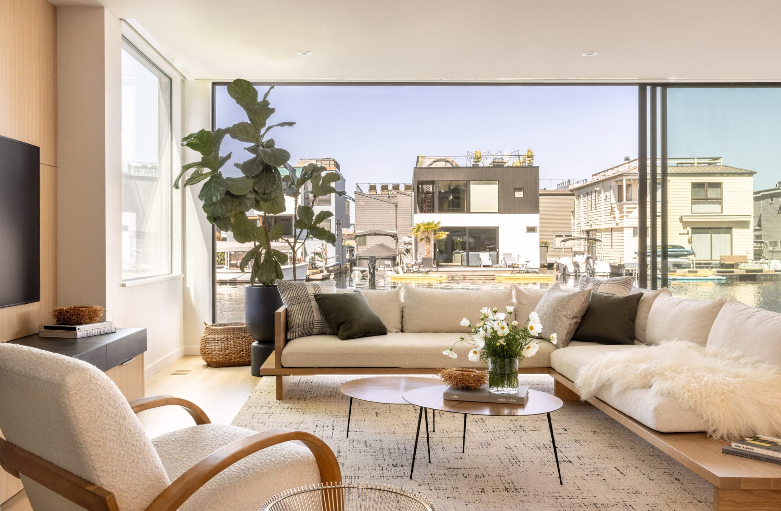 Modern living room with large windows, a beige sofa, a white armchair, a circular coffee table, and a potted plant. Outdoor view of buildings and water is visible through the windows.