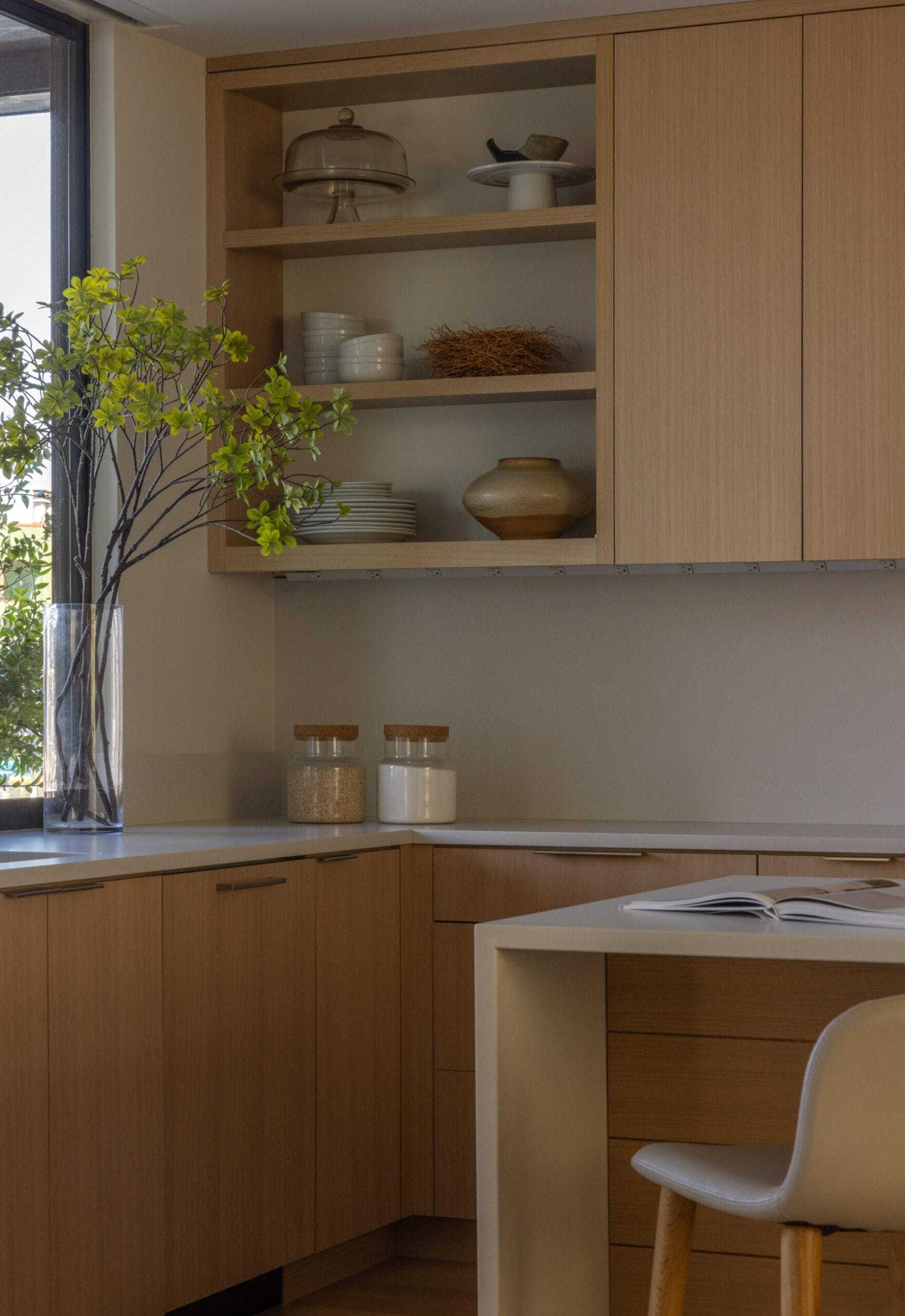 Modern kitchen corner with light wooden cabinets, open shelving displaying dishes, a vase with branches, two jars, and a chair by a table with an open book. Natural light from the window.