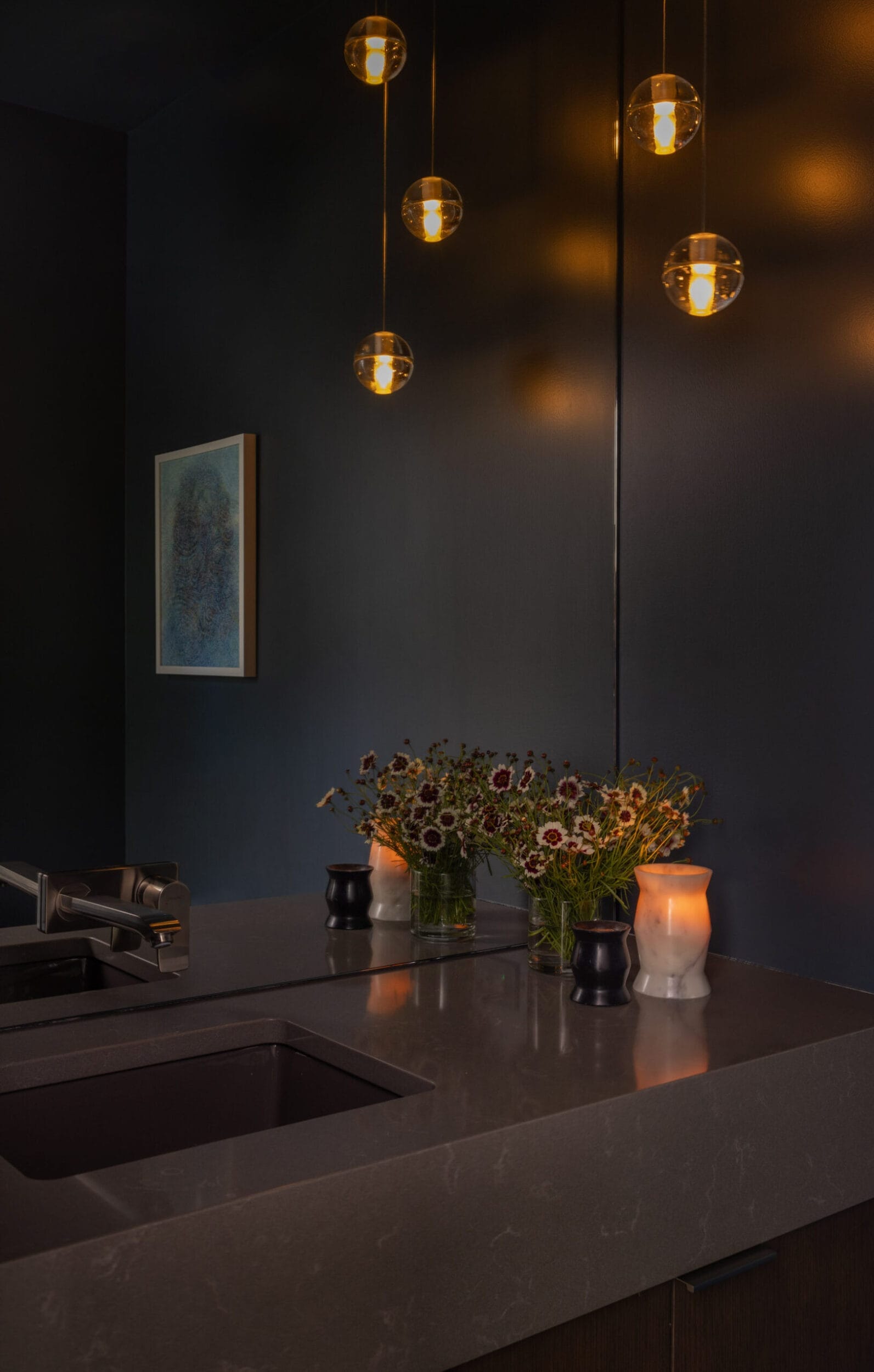 A dimly lit bathroom with hanging pendant lights, a modern sink, flowers in vases, a lit candle, and a picture on the dark wall.