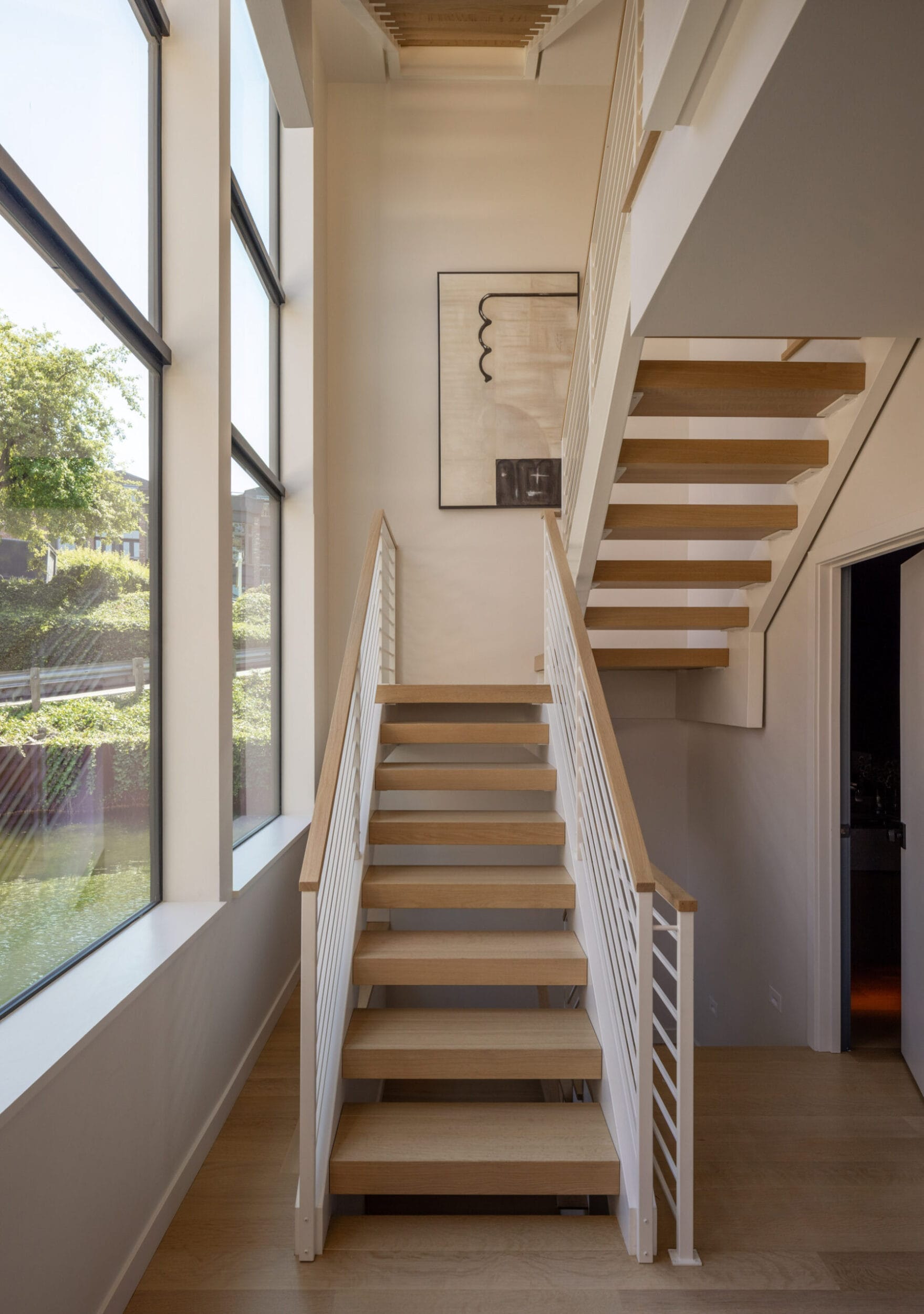 Modern interior staircase with wooden steps and white railings, leading to an upper level. Large windows on the left side provide natural light. Minimalist artwork hangs on the wall.