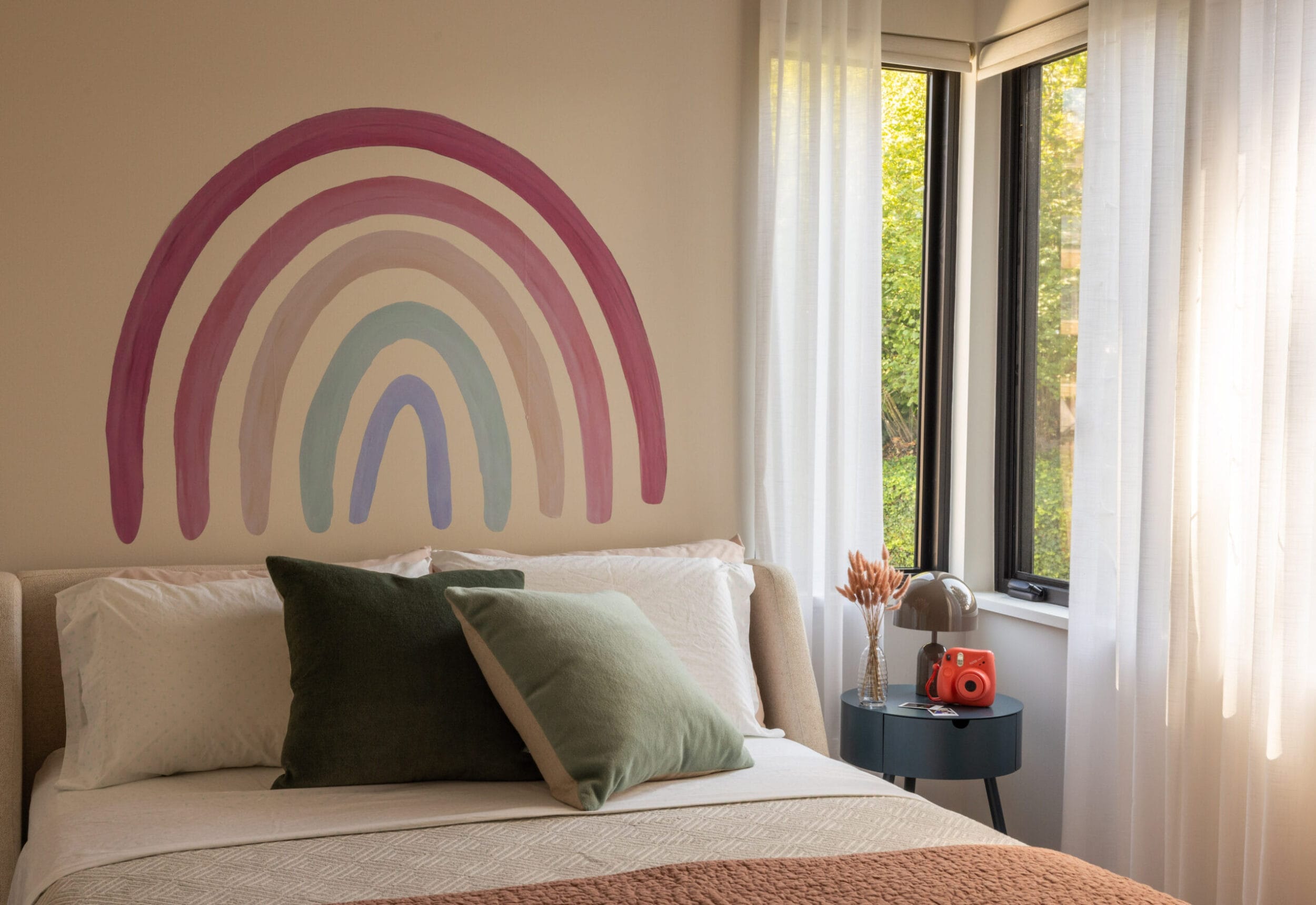 Bedroom with a rainbow mural on the wall, a bed with green and brown pillows, a table with a toy camera, and a lamp by a window with white curtains.