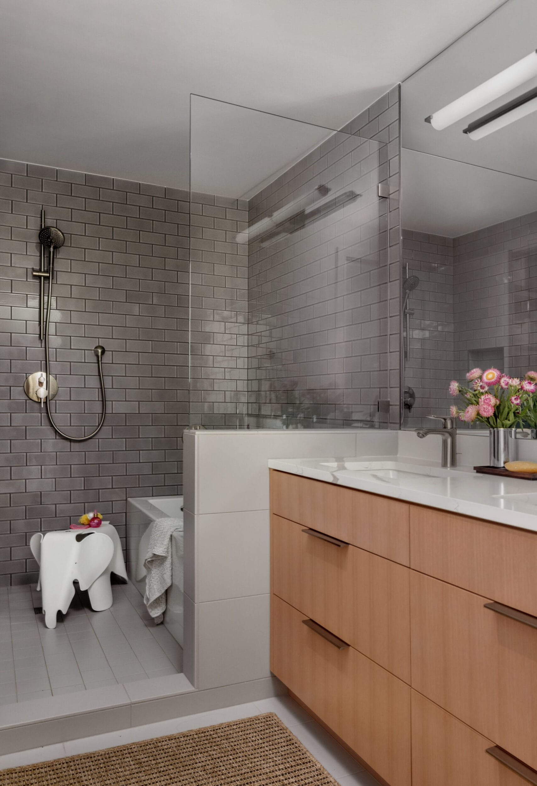 Modern bathroom with a glass-enclosed shower, gray subway tile, wood vanity with dual sinks, and a decorative stool. A vase with flowers is placed on the countertop.