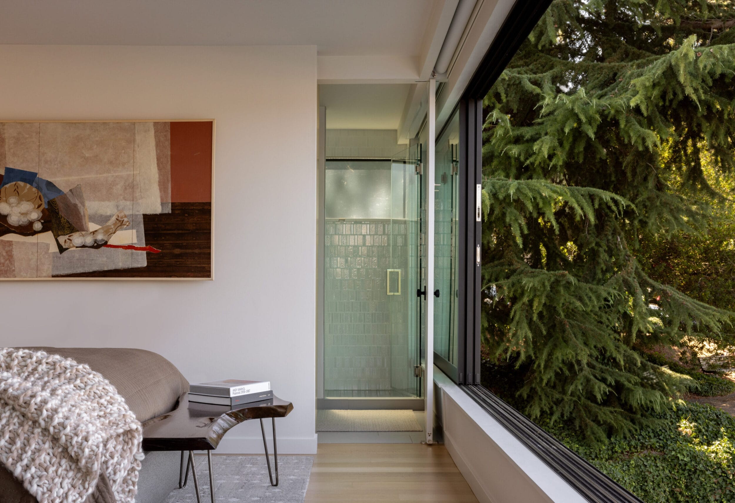 Modern room with a couch, textured blanket, books, large artwork, glass shower, and floor-to-ceiling window overlooking greenery.