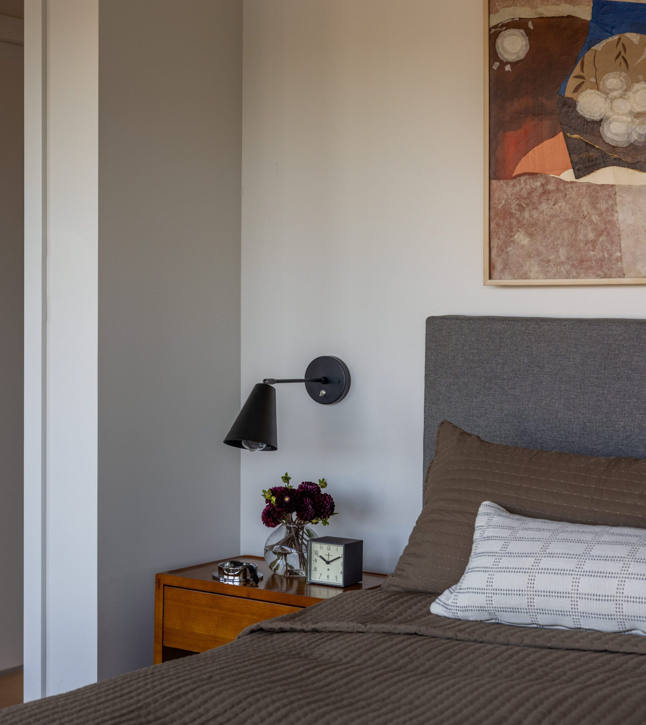 A neatly made bed with a gray headboard and brown bedding is beside a wooden nightstand with a black lamp, a clock, and flowers. A painting hangs on the wall above.