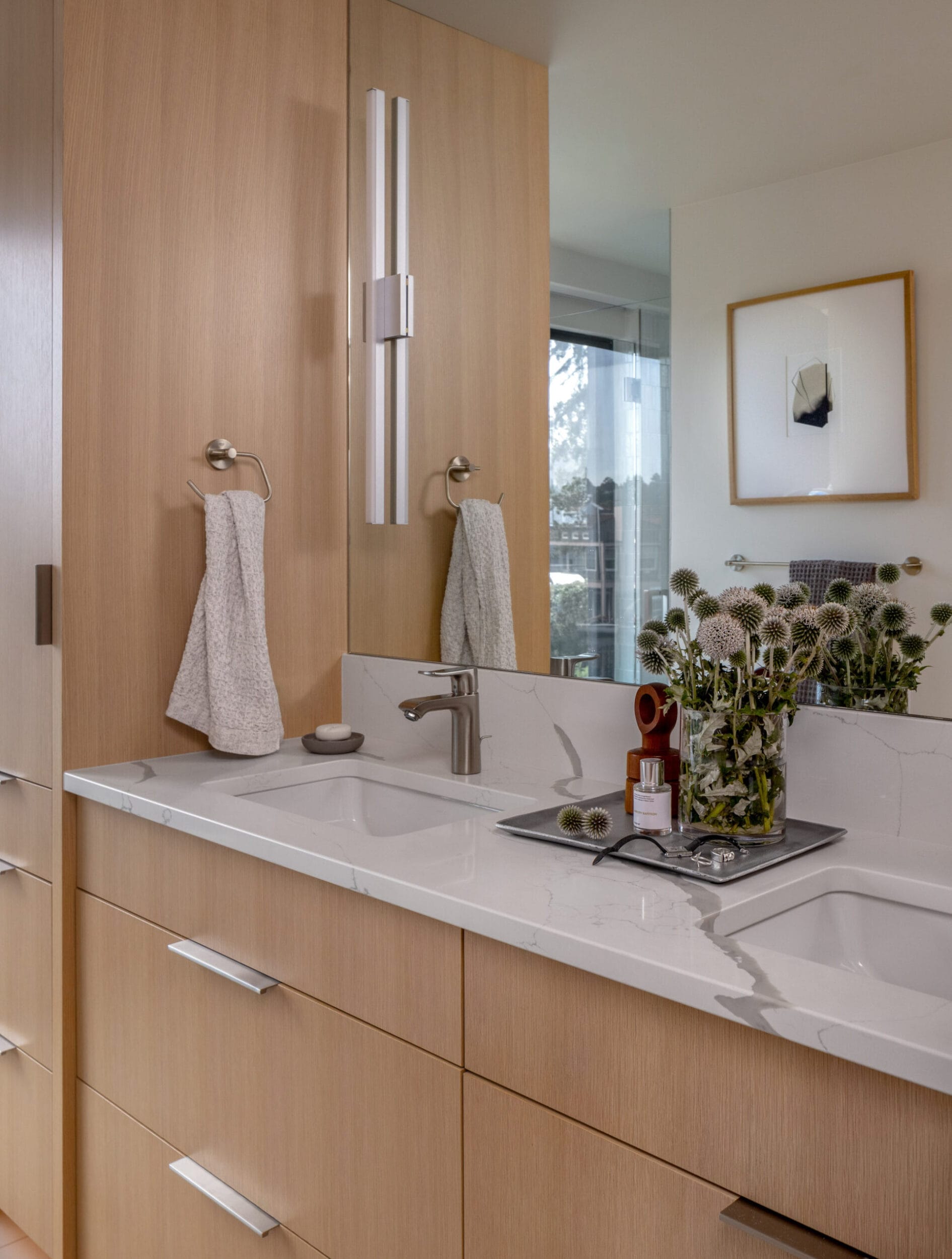 A modern bathroom features a wooden cabinet, marble countertop with two sinks, a tall mirror, and a vase of flowers. A towel hangs on the wall and a small painting is visible in the background.